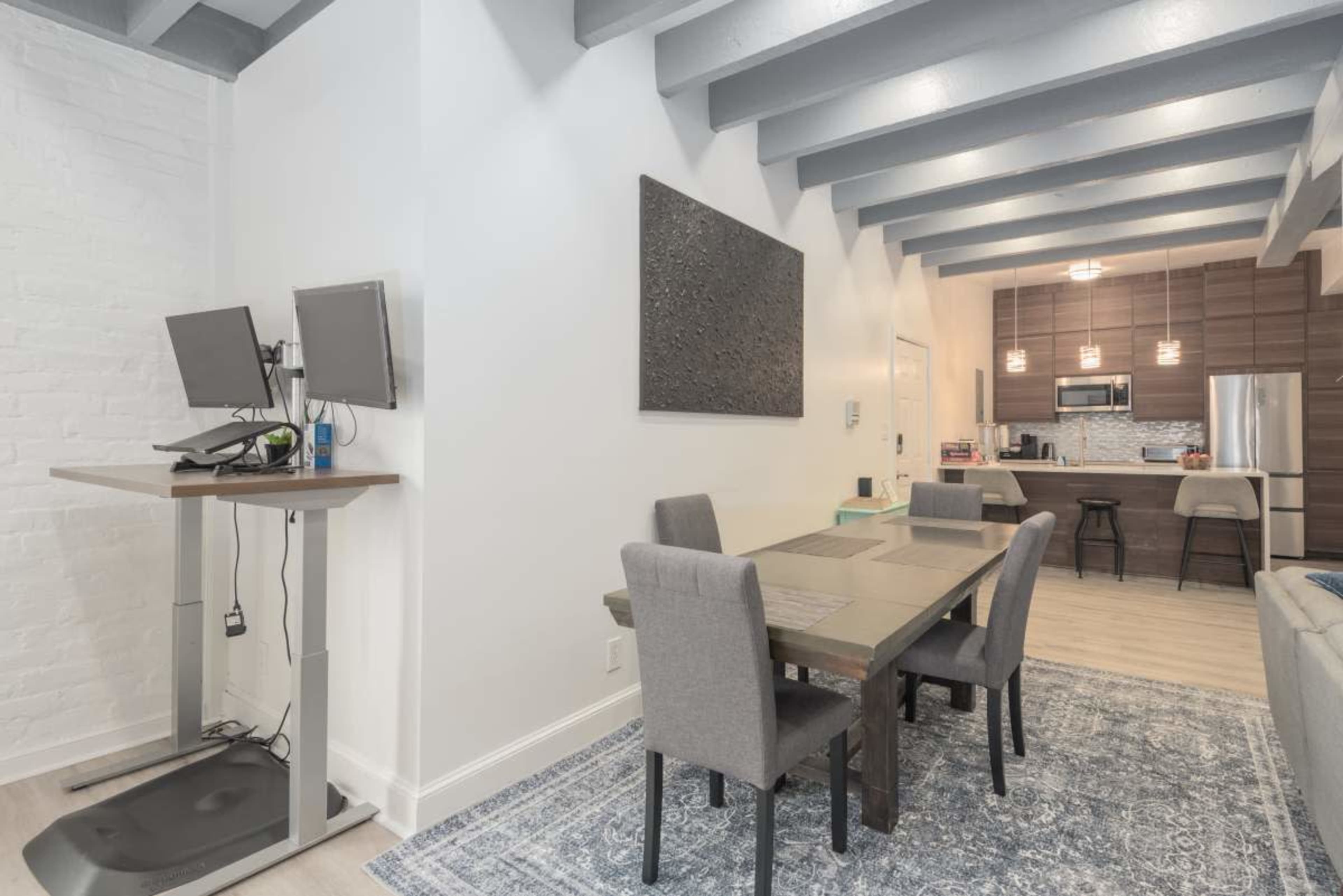 The image shows a modern dining area with a table and chairs, leading to a kitchen visible in the background.