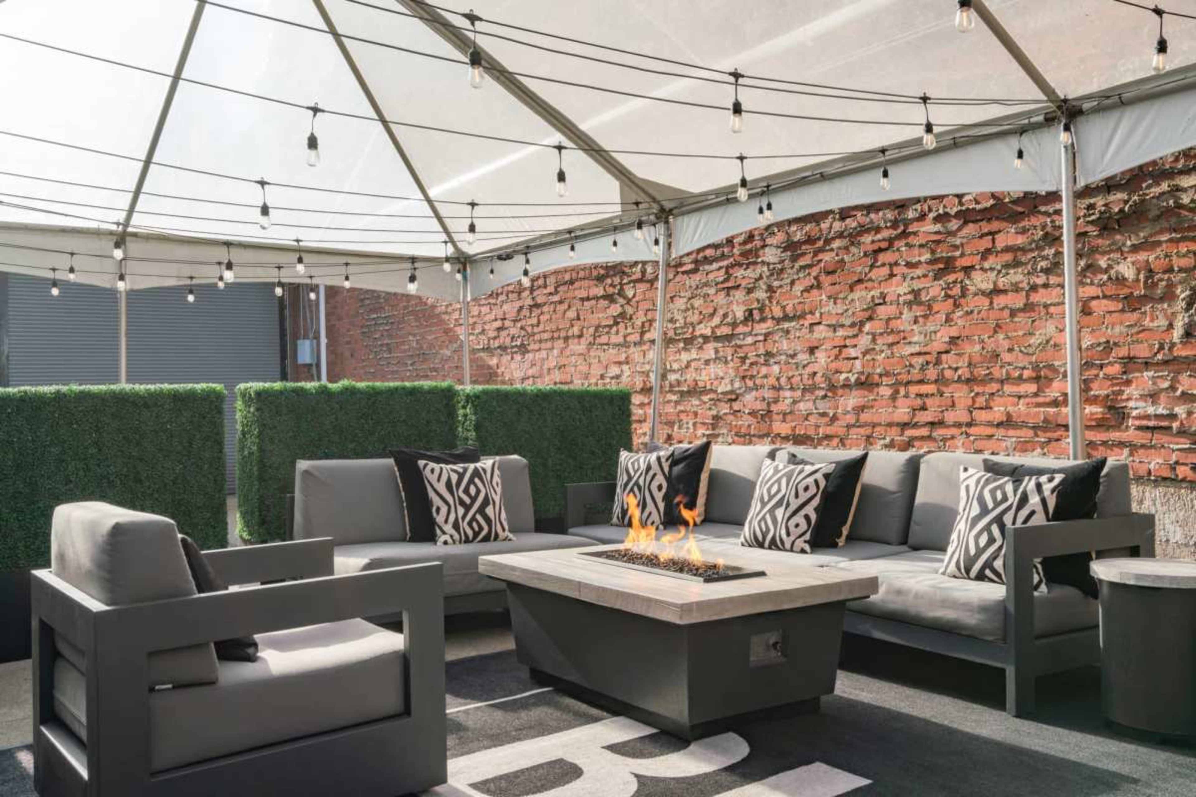 A covered outdoor seating area features a sectional sofa with decorative pillows surrounding a gas fire pit, all set against a brick wall and green hedge.