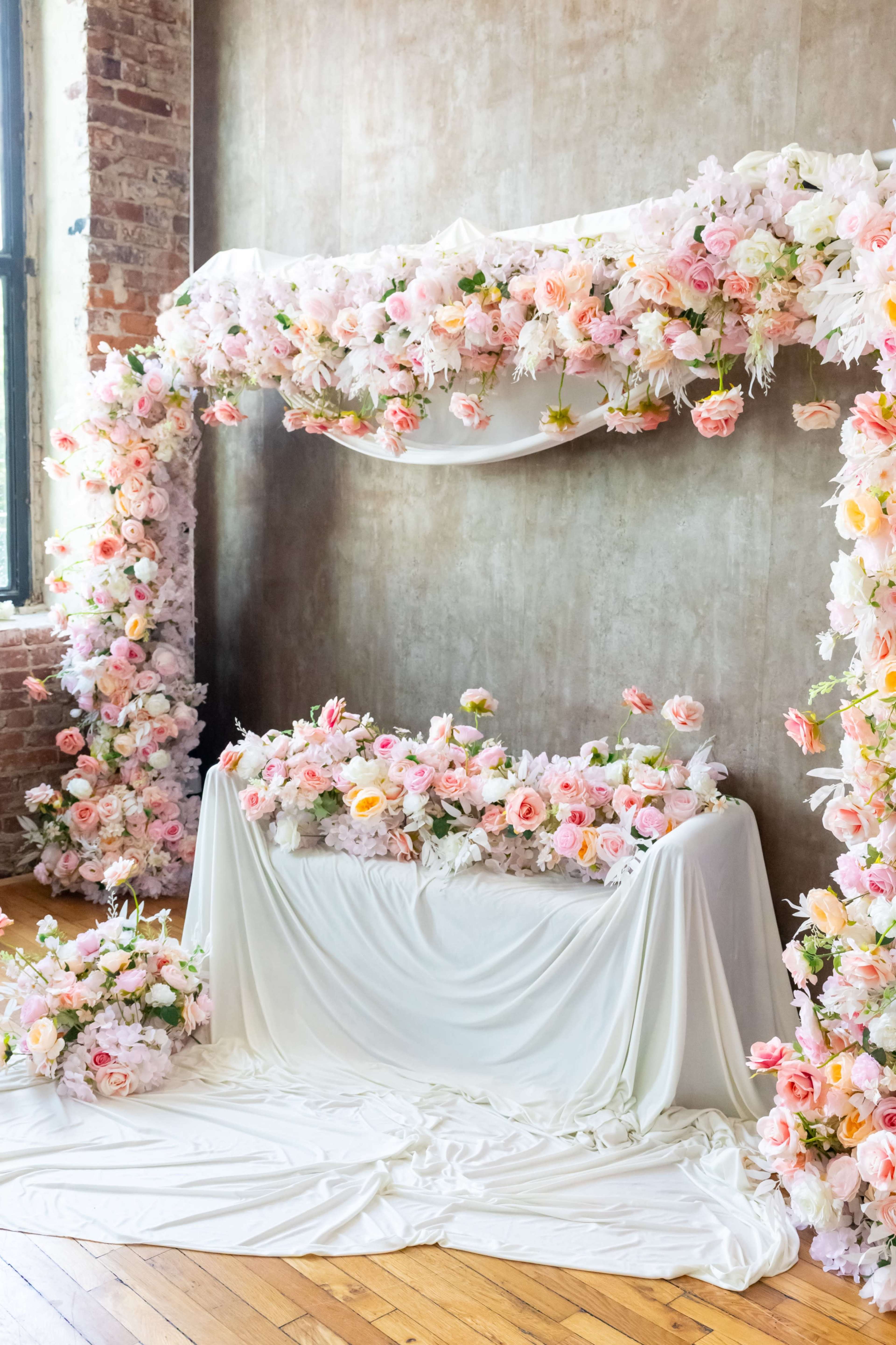 The image shows a floral display arch adorned with pink and white flowers, draped fabric, and a matching floral arrangement on a table in front, set against a textured wall and wooden floor.