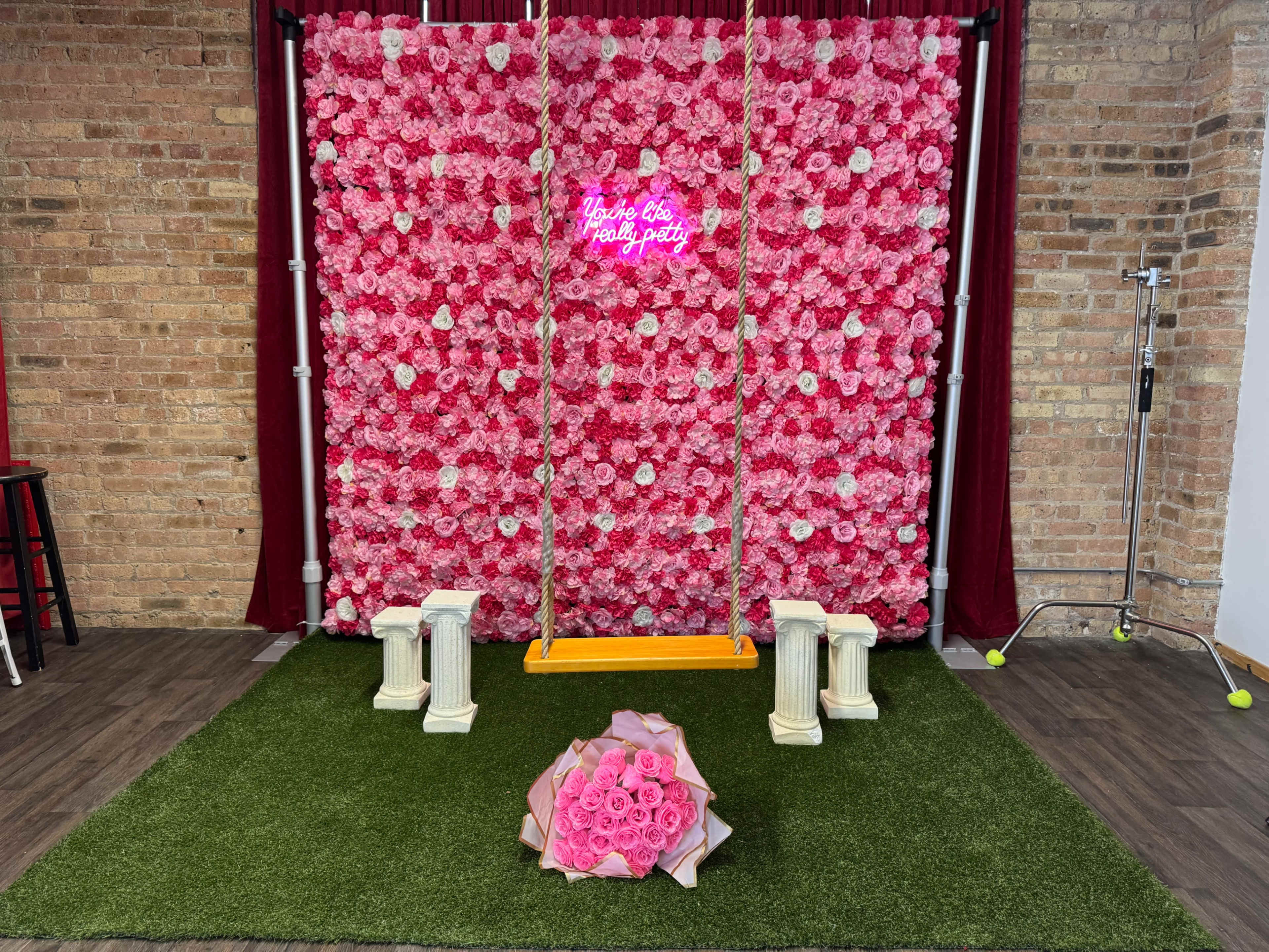 The image shows a vibrant backdrop of pink flowers with a swing in front, adorned with a bouquet of roses and artificial grass flooring.