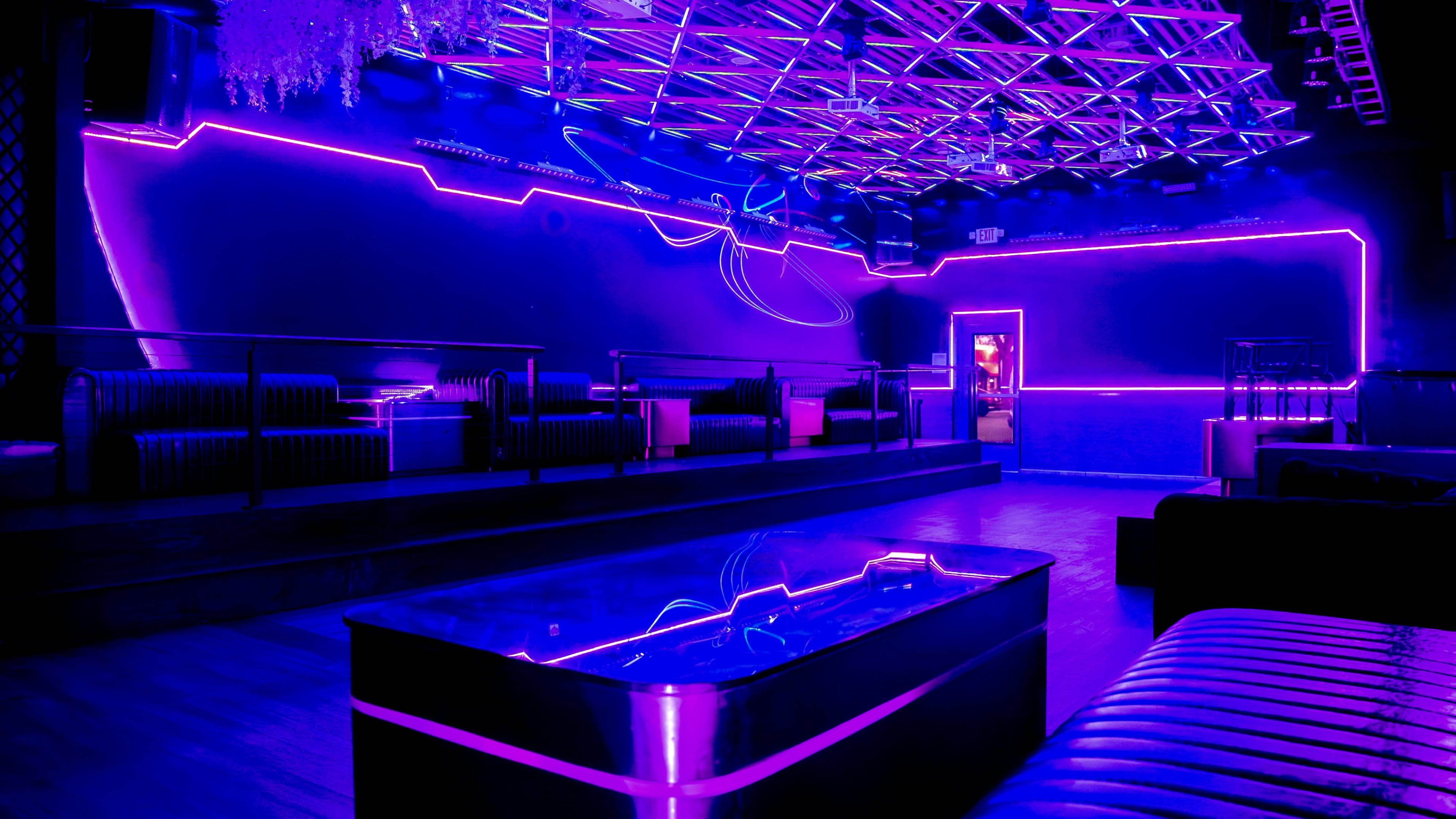 The image shows an empty nightclub interior featuring purple neon lighting and modern seating arrangements.