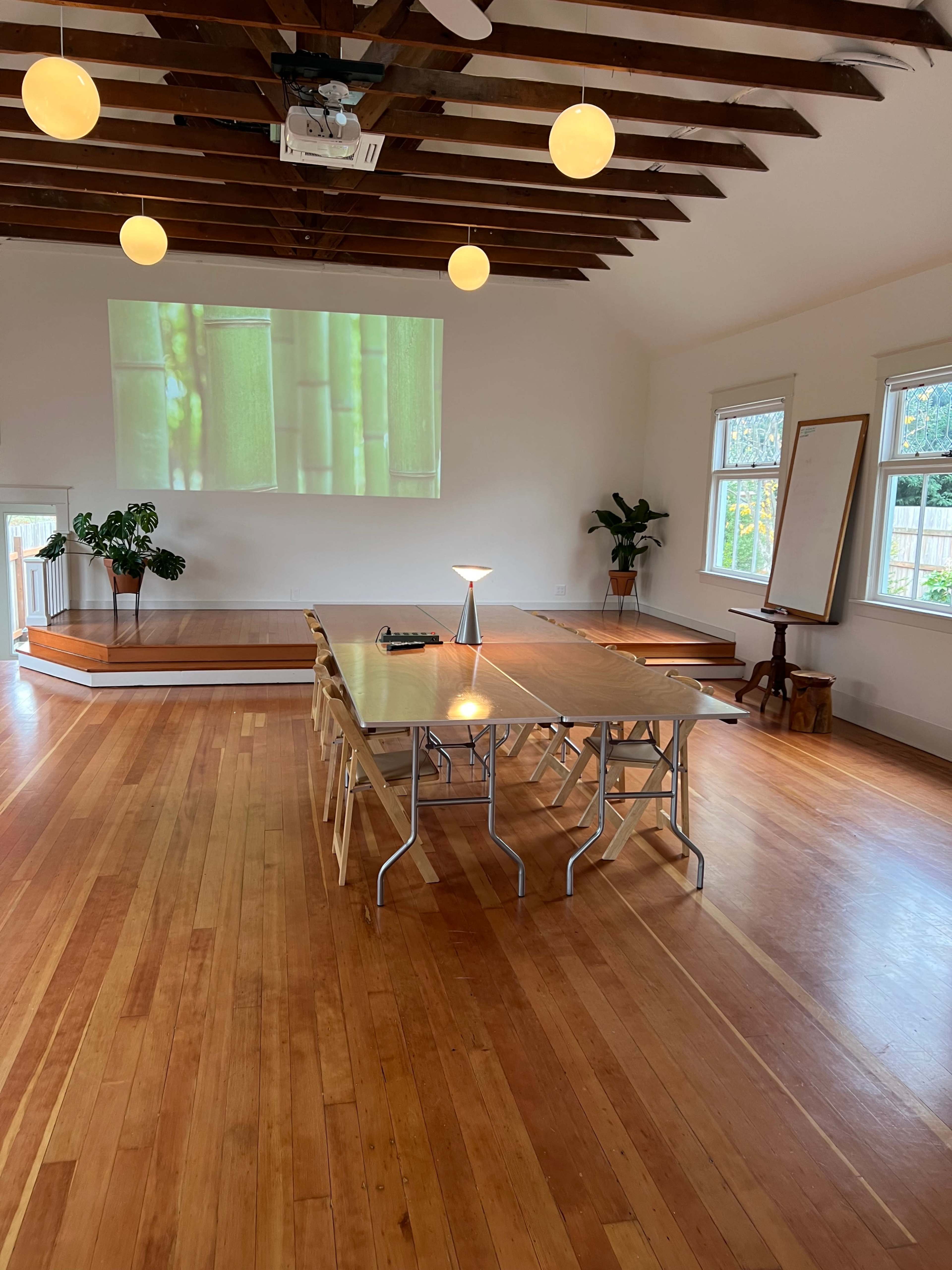 The image shows a spacious, well-lit room with wooden floors, featuring a large table set for a meeting, a projector displaying bamboo on the wall, and potted plants in the corners.