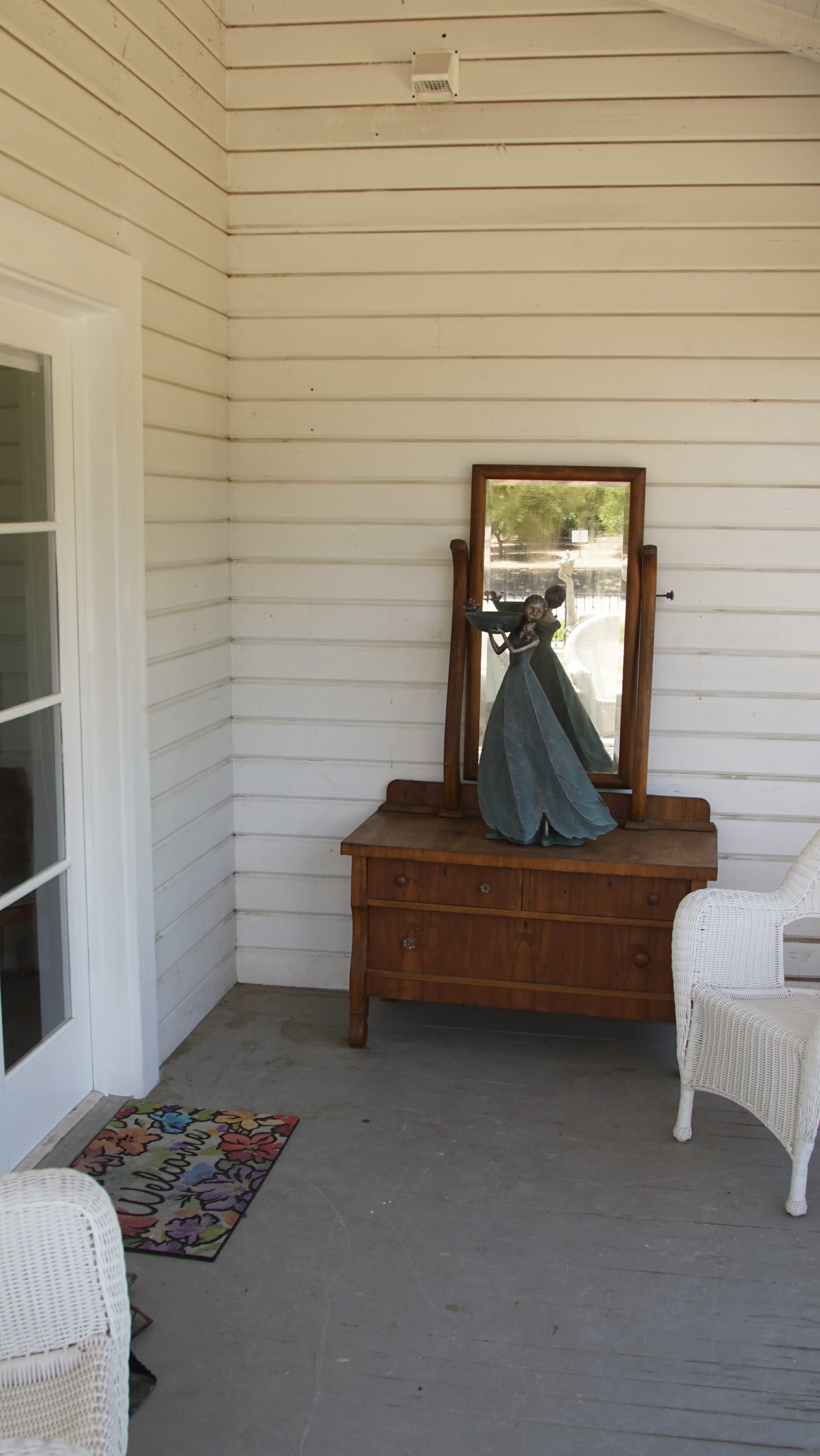 A wooden dresser with a mirror displays a dress form wearing a blue garment, situated in a porch space beside a white door and wicker chairs.