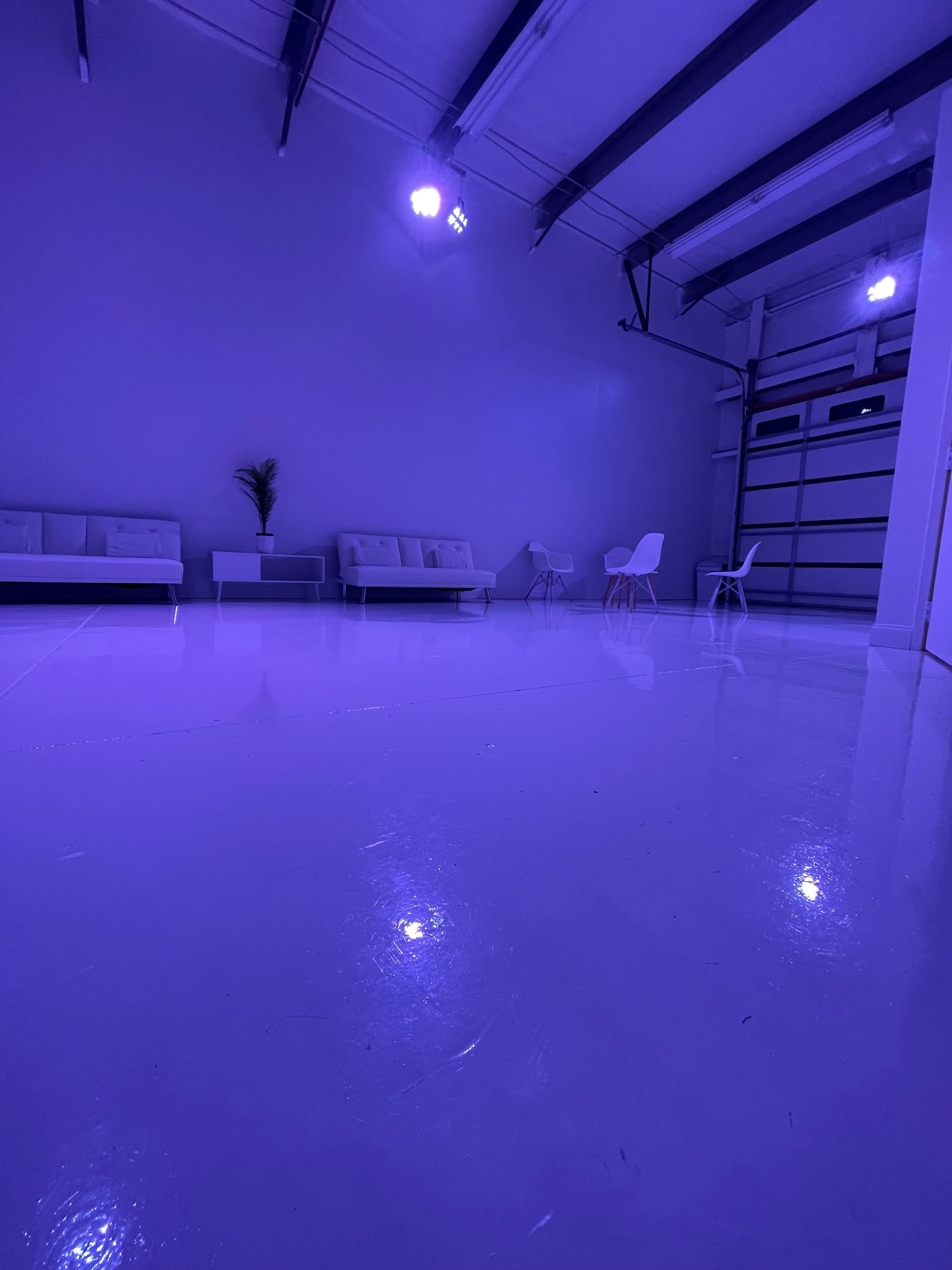 A spacious, empty room with a glossy white floor, a few white couches, chairs, and purple lighting.