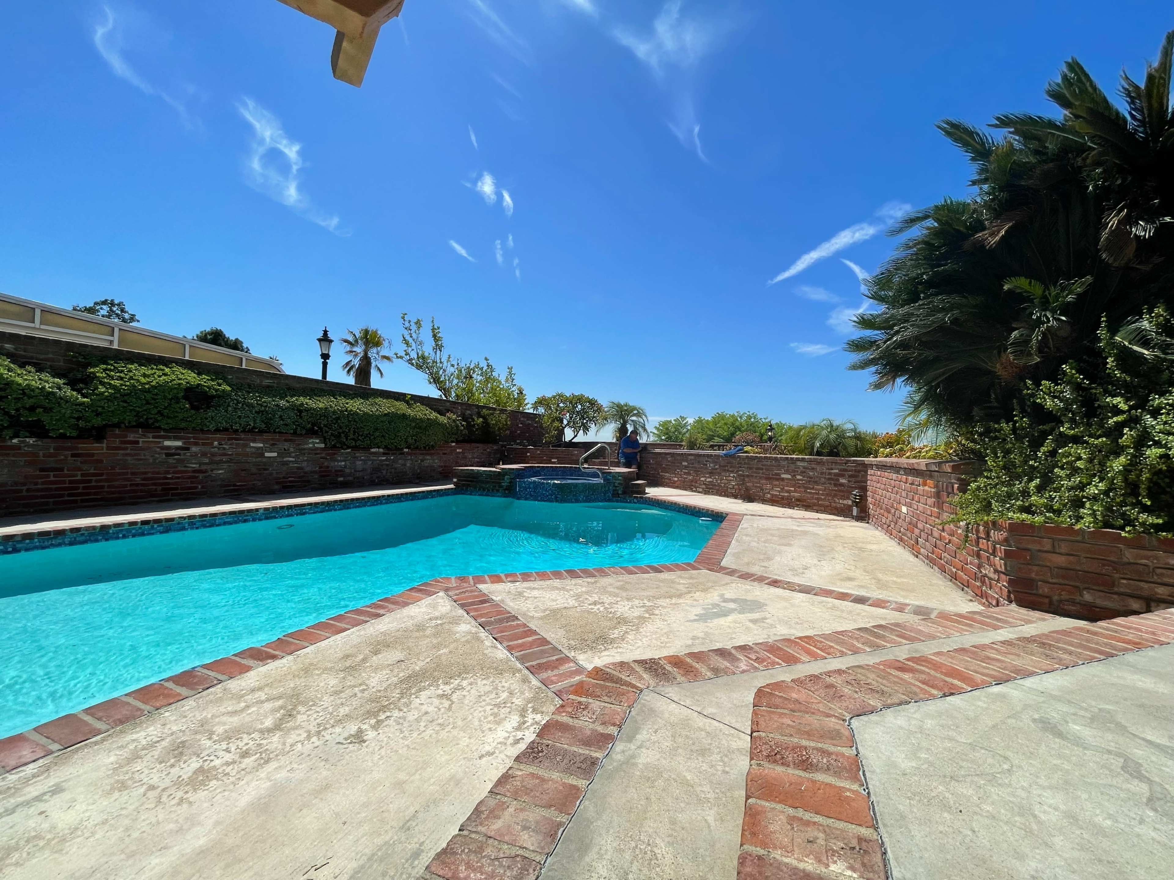 A clear blue swimming pool surrounded by brick patios and lush greenery under a sunny sky.