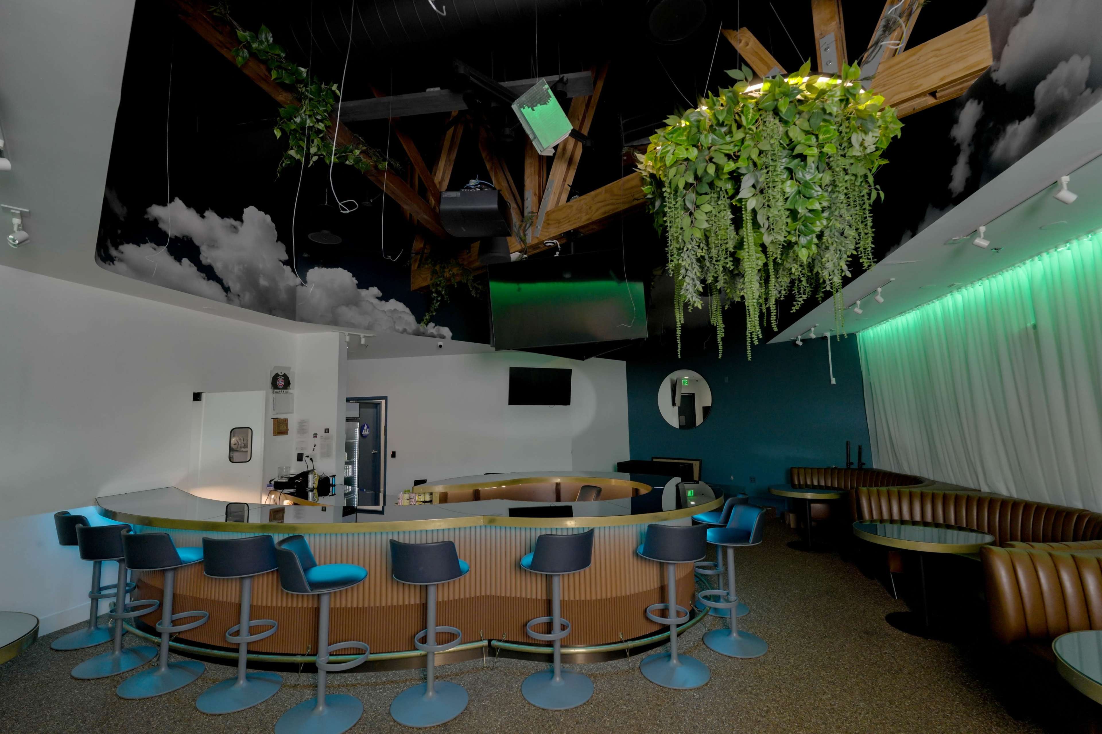 The image features a modern bar interior with a curved counter, high stools, and greenery hanging from the ceiling, surrounded by semi-circular booths against the walls.