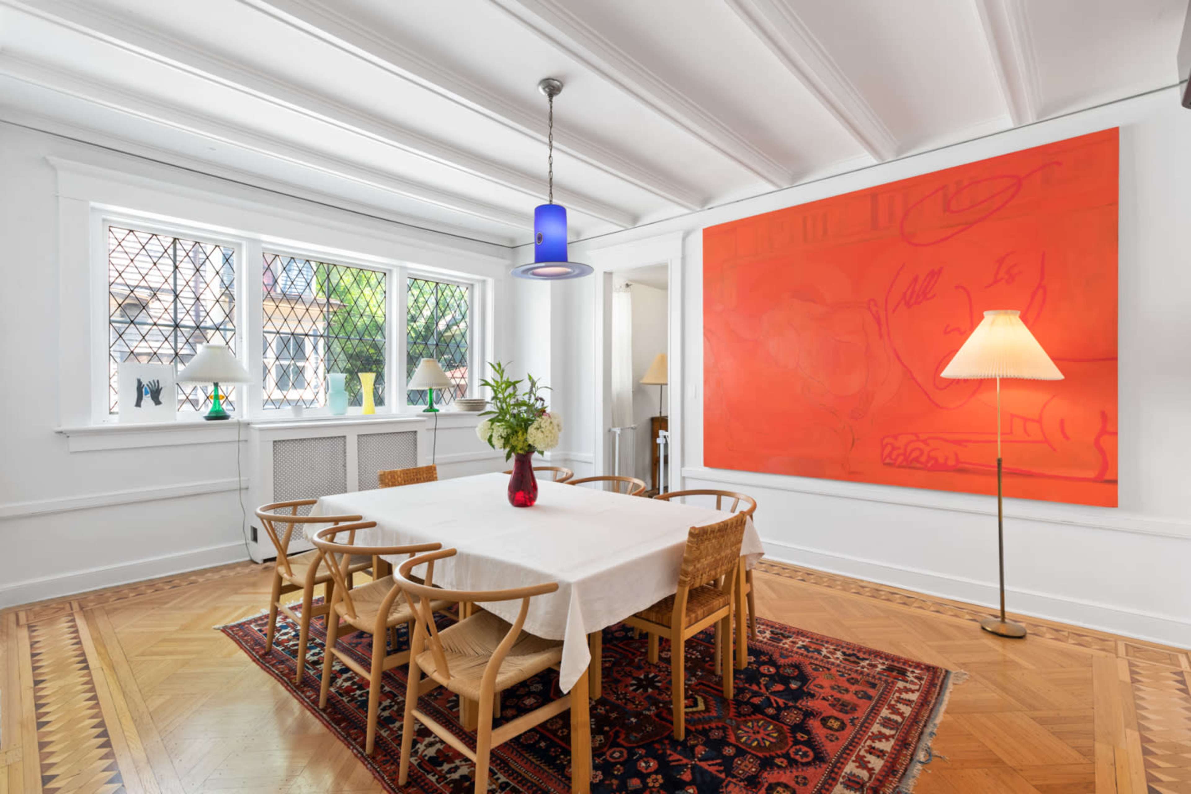 The image shows a dining area with a table surrounded by chairs, a large orange artwork on the wall, and a floor lamp beside the table.