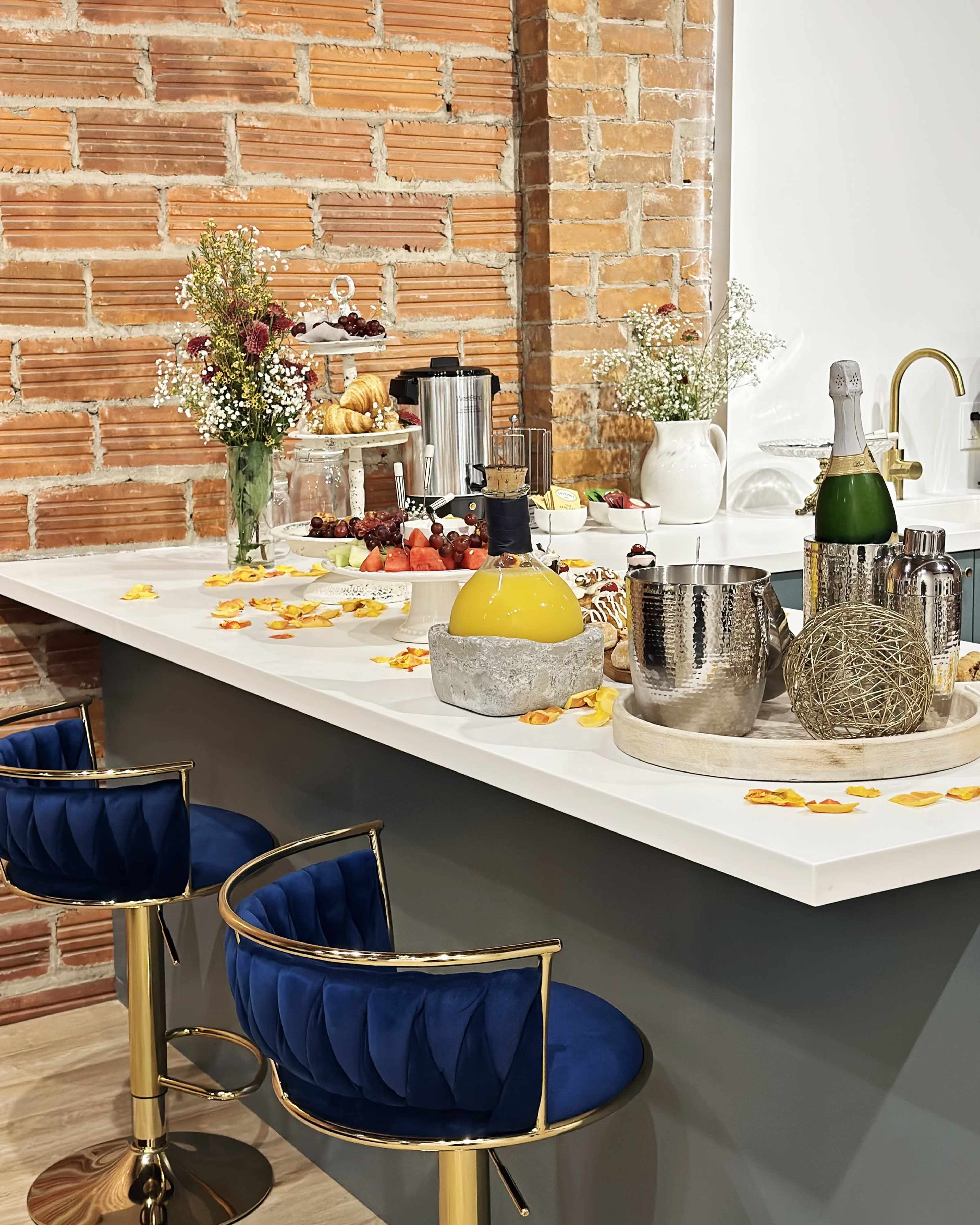 A modern kitchen island is adorned with various food items, a bouquet of flowers, and decorative elements, set against a backdrop of exposed brick walls.