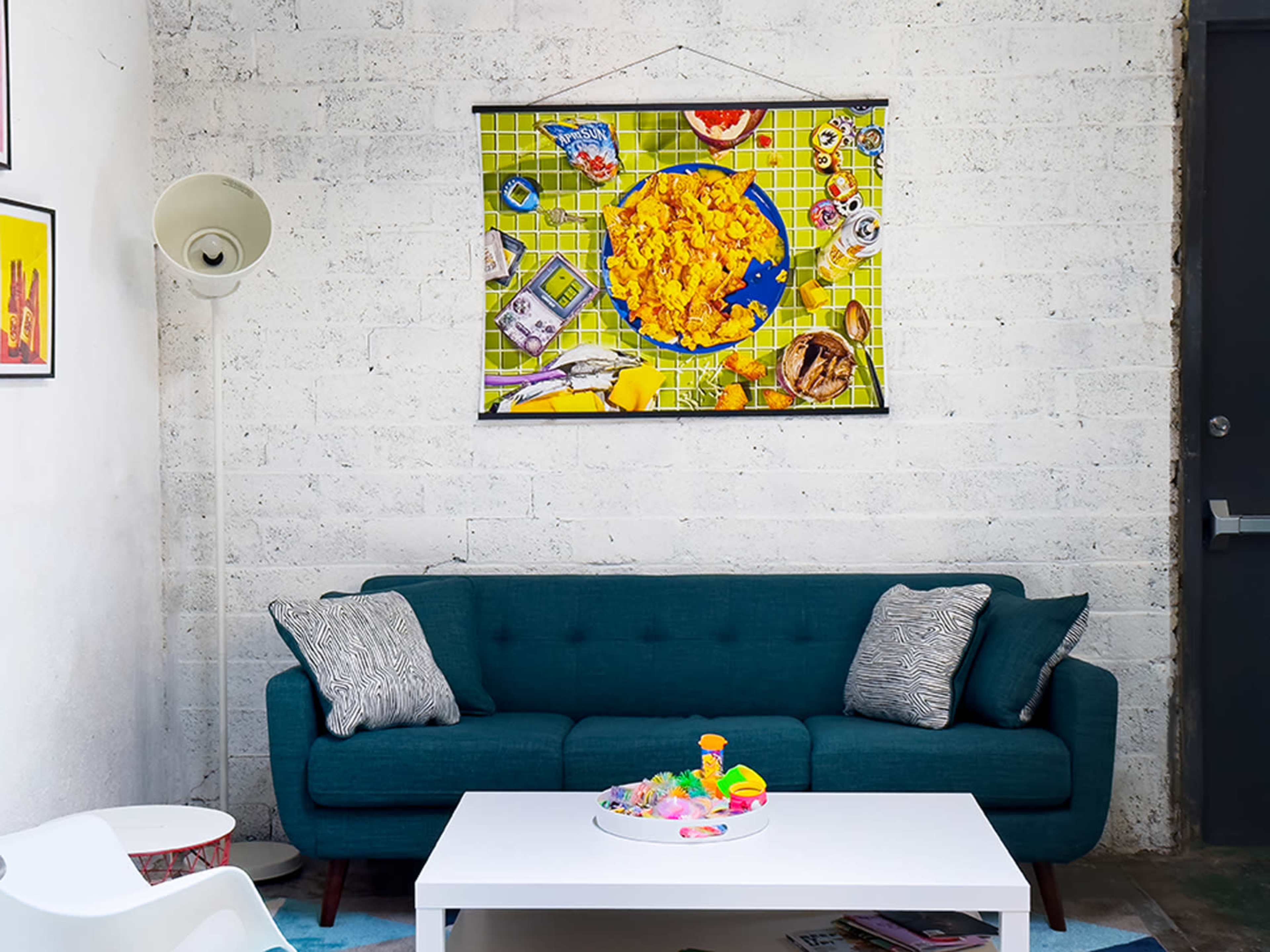 The image shows a modern living room with a teal sofa, a white coffee table with colorful objects, and a bright wall hanging above the sofa.