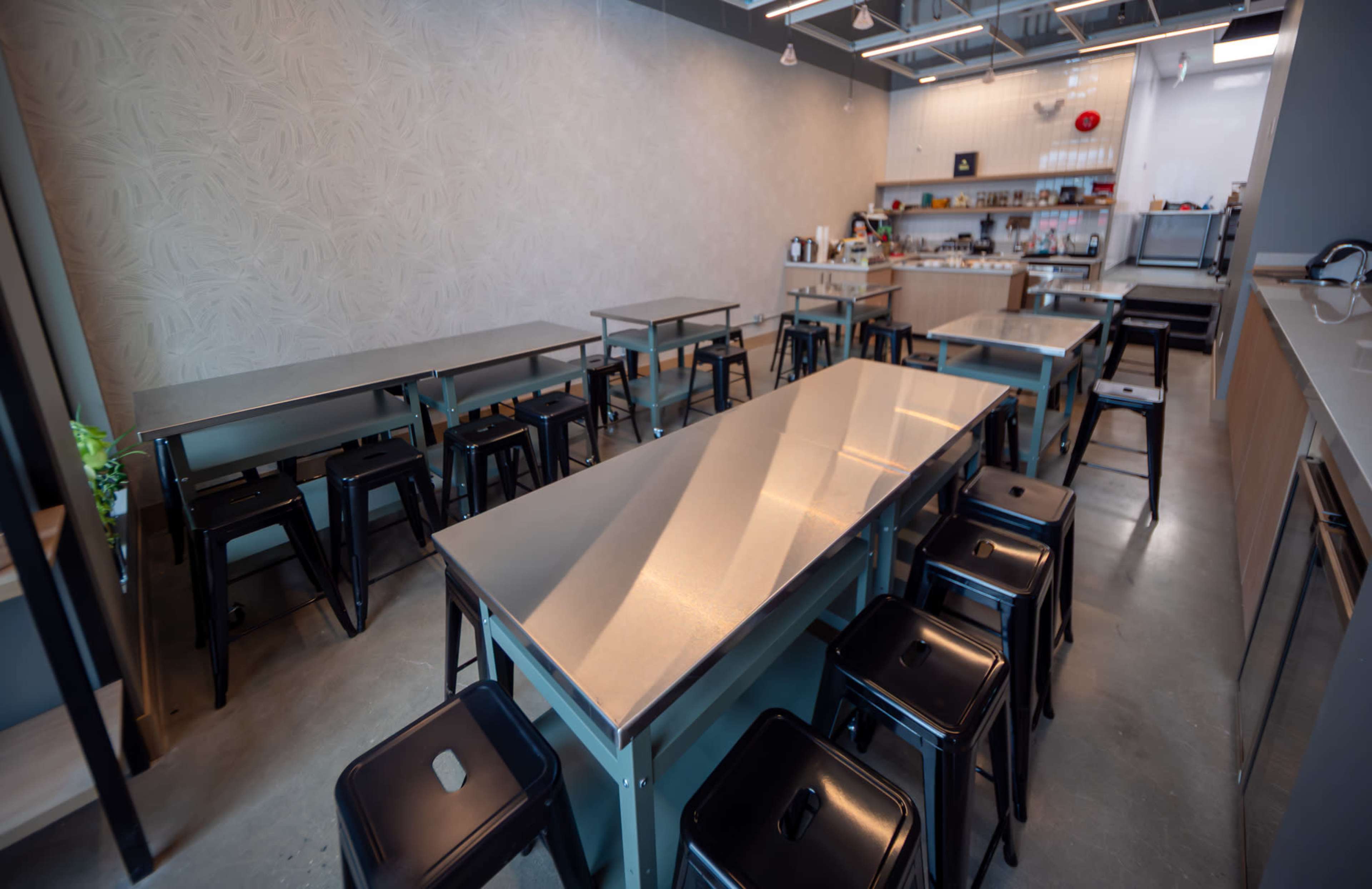 The image shows a spacious, modern cafe interior with several steel tables and black stools arranged neatly.