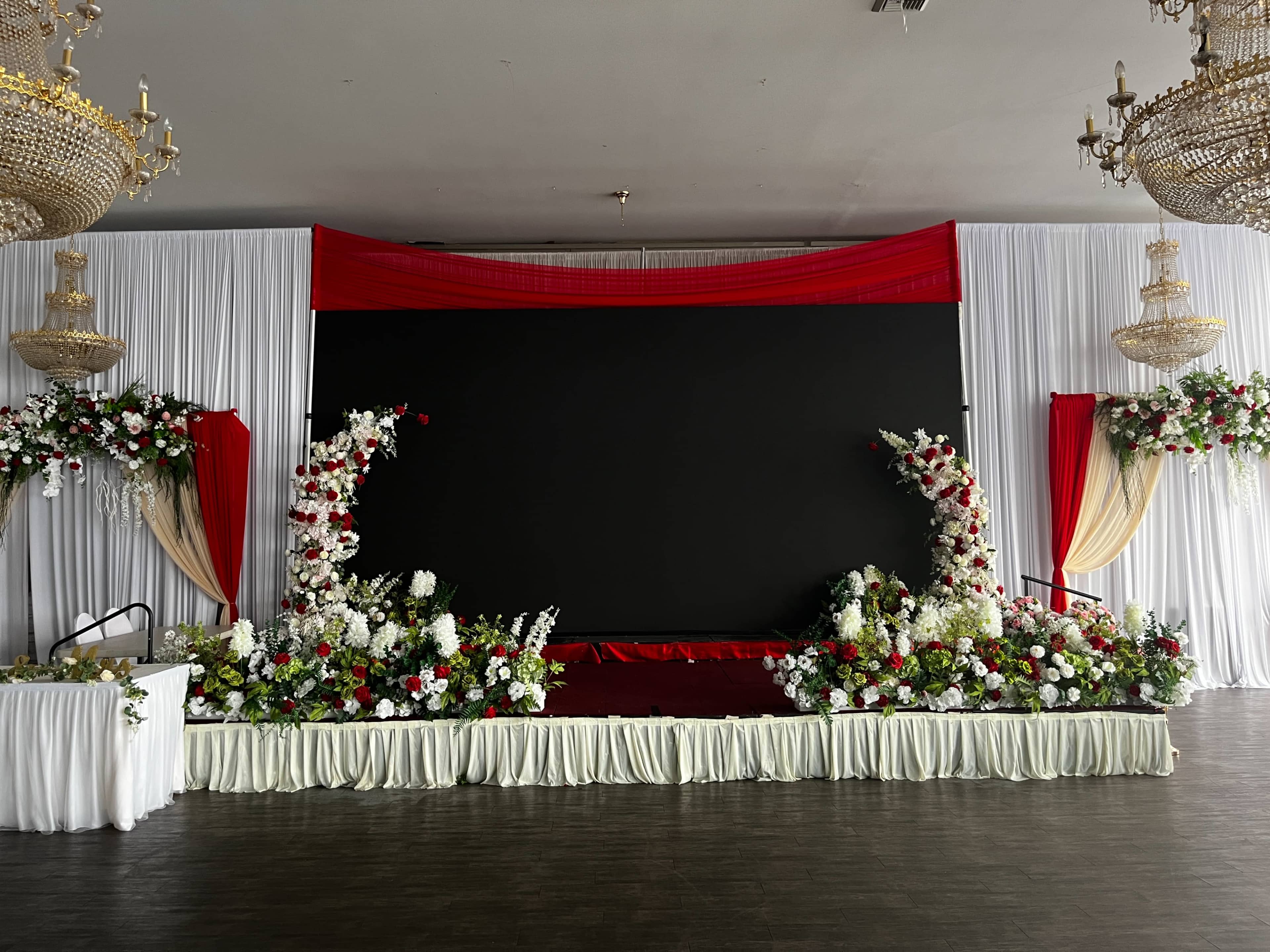 The image shows a decorated stage with a large black screen, surrounded by floral arrangements and draped fabric in white and red.