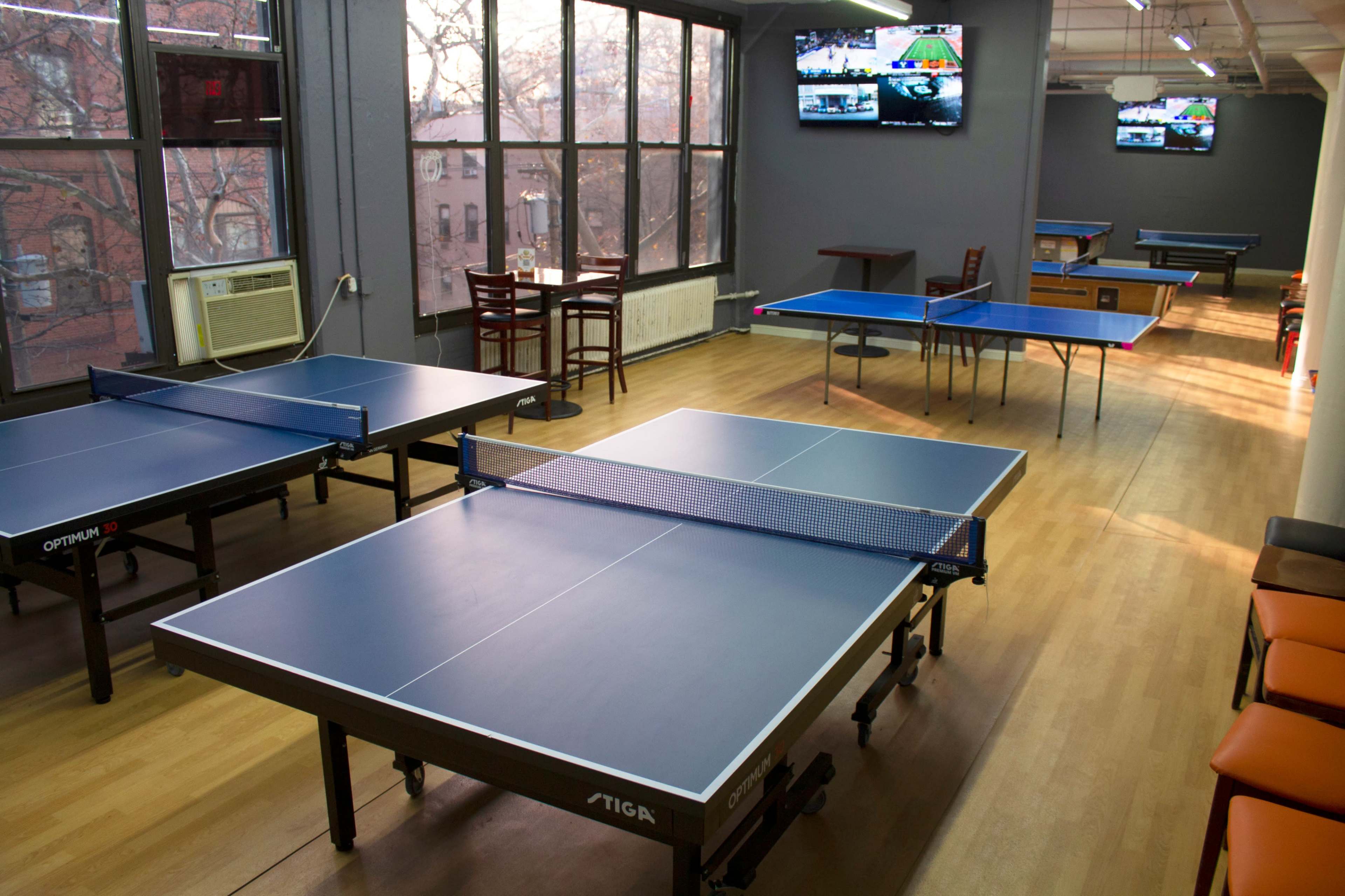 The image shows a table tennis facility featuring several blue tables, an air conditioning unit, and multiple wall-mounted screens displaying games.