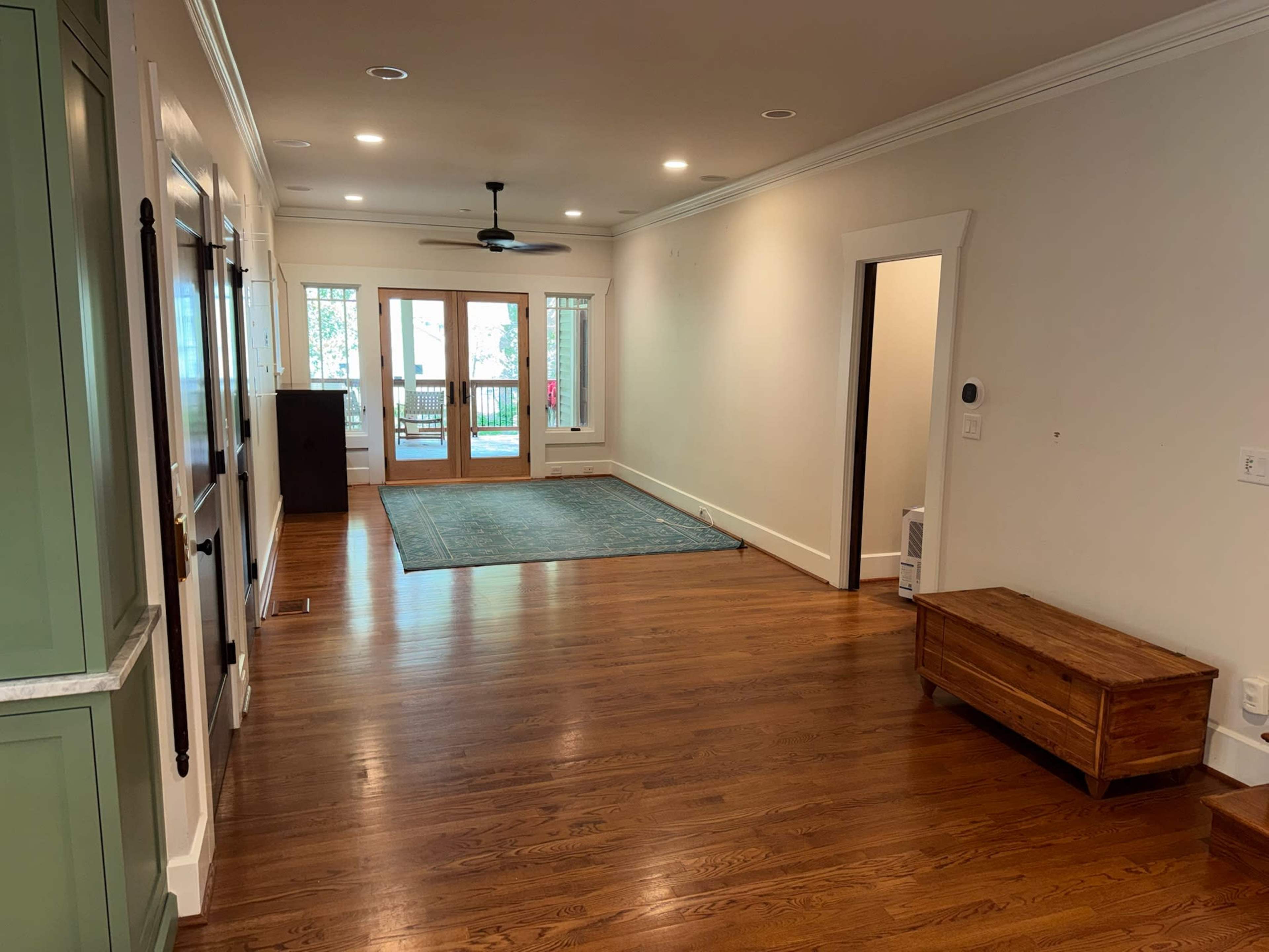 The image shows a spacious, well-lit interior of a room with wooden flooring, a ceiling fan, and a doorway leading to a balcony.