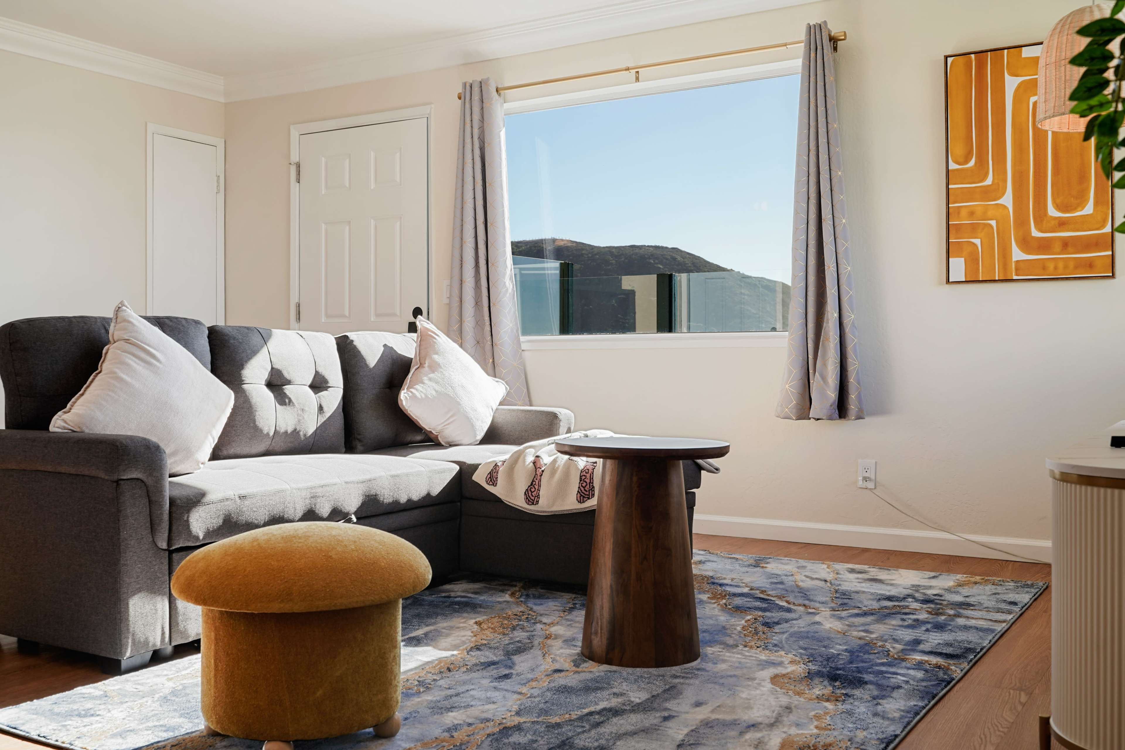 A gray sofa with decorative pillows is positioned in a light-filled living room, featuring a round wooden table and a yellow ottoman on a patterned rug.