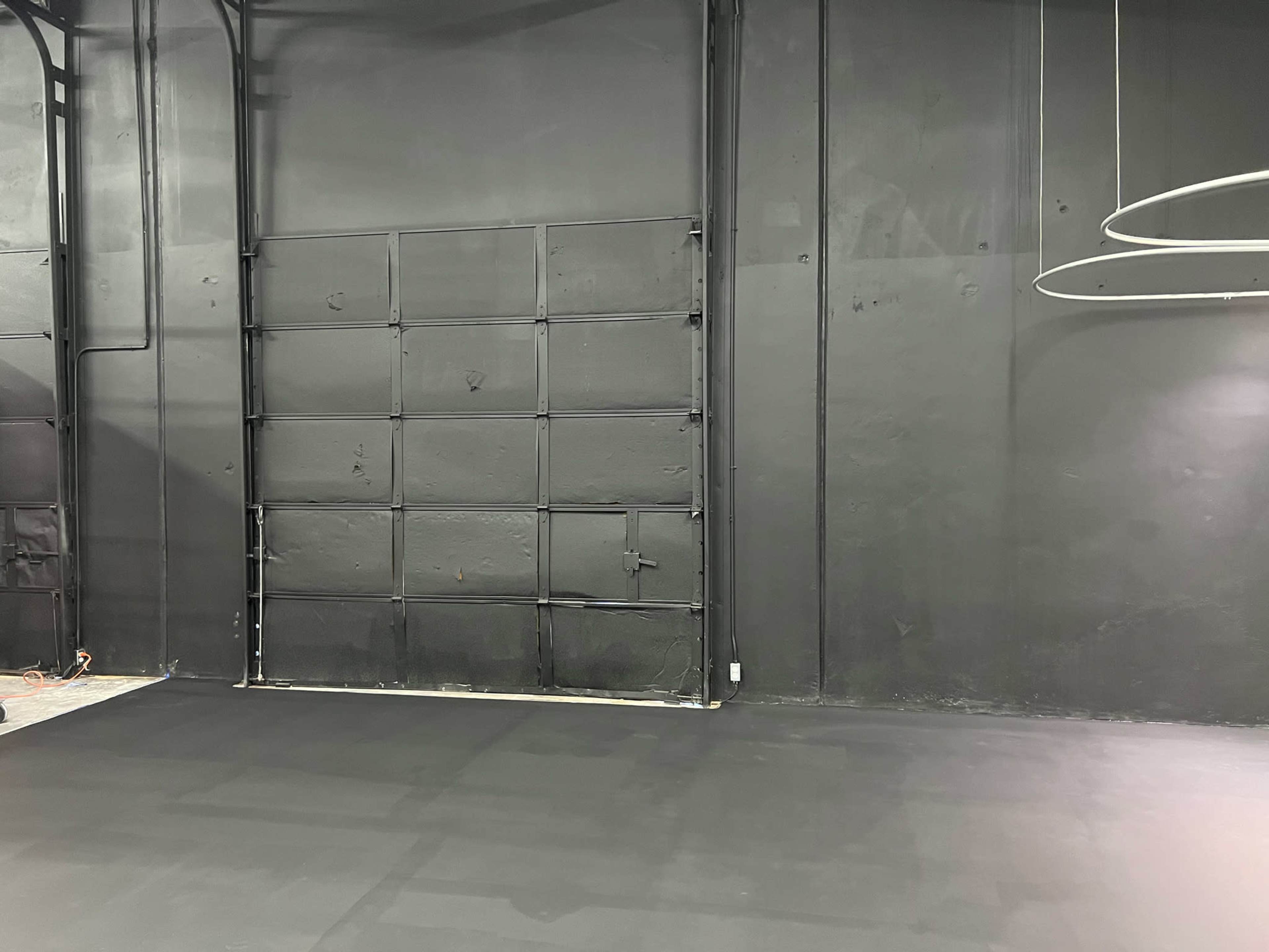 A dimly lit room with a flat black floor and a large black wall featuring a closed metal garage door.