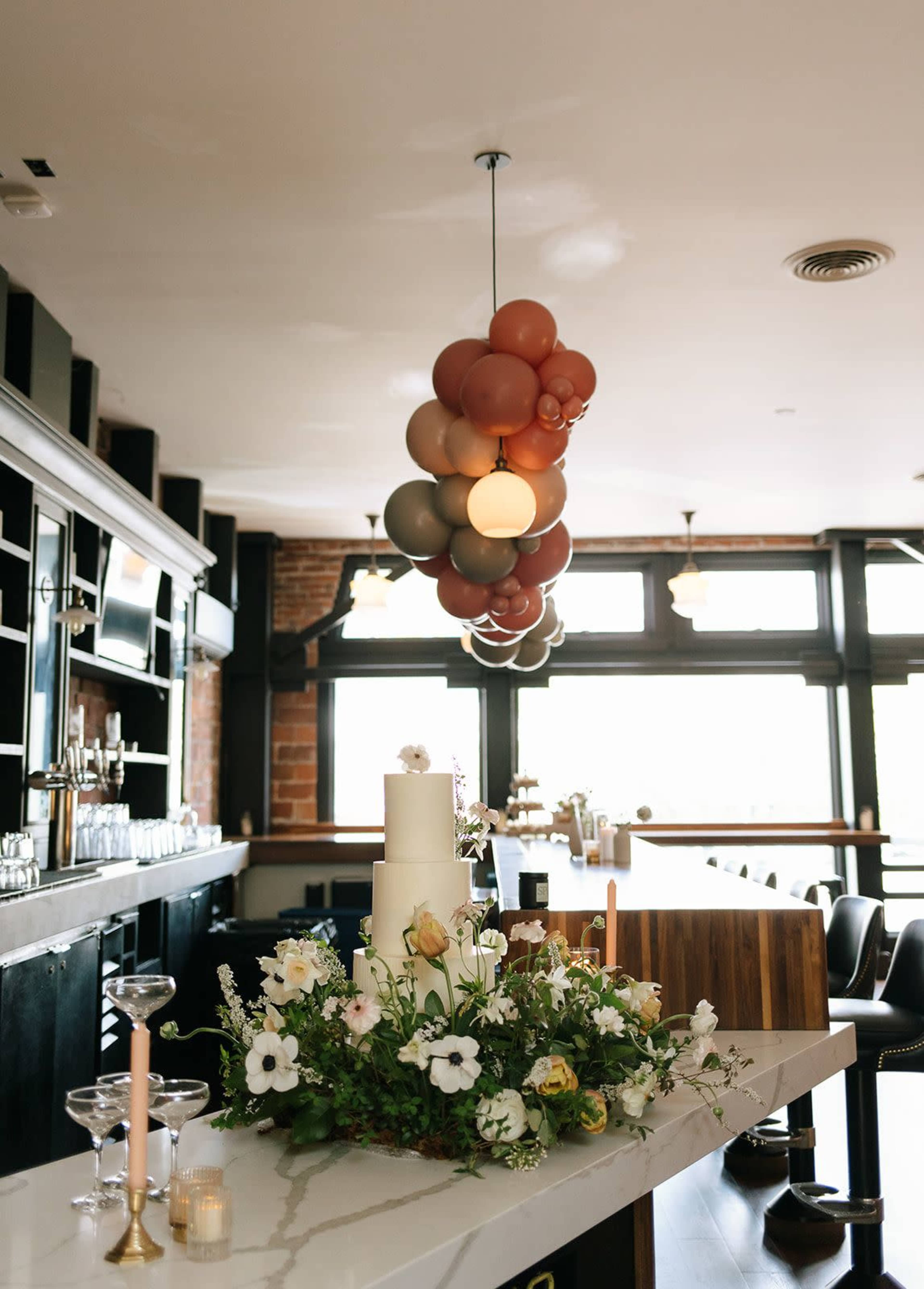 A modern bar setting features a tiered white cake adorned with flowers, complemented by a colorful balloon arrangement overhead.