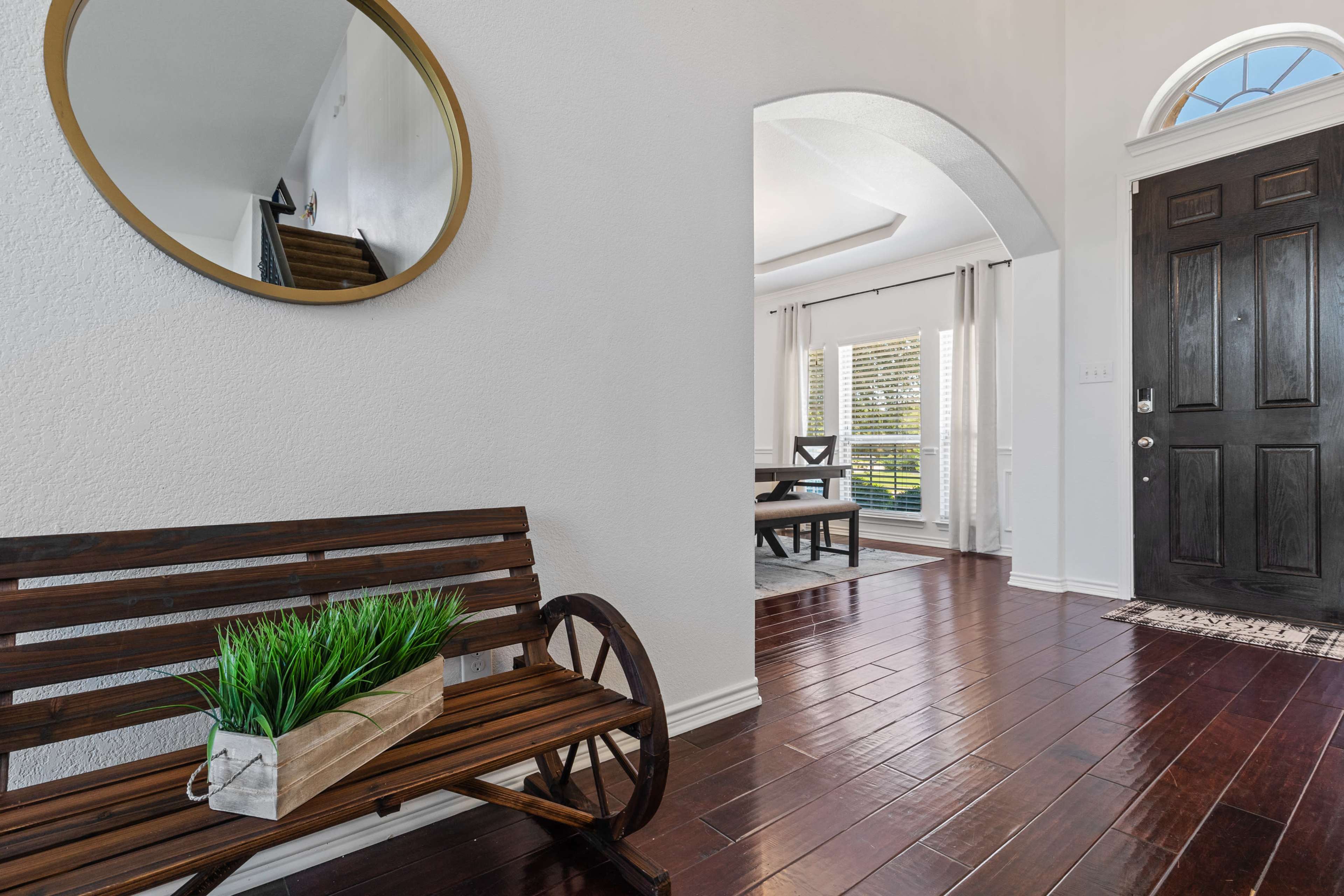 The entrance hall features a wooden bench with a planter, a large round mirror, and a view of a dining area through an archway.