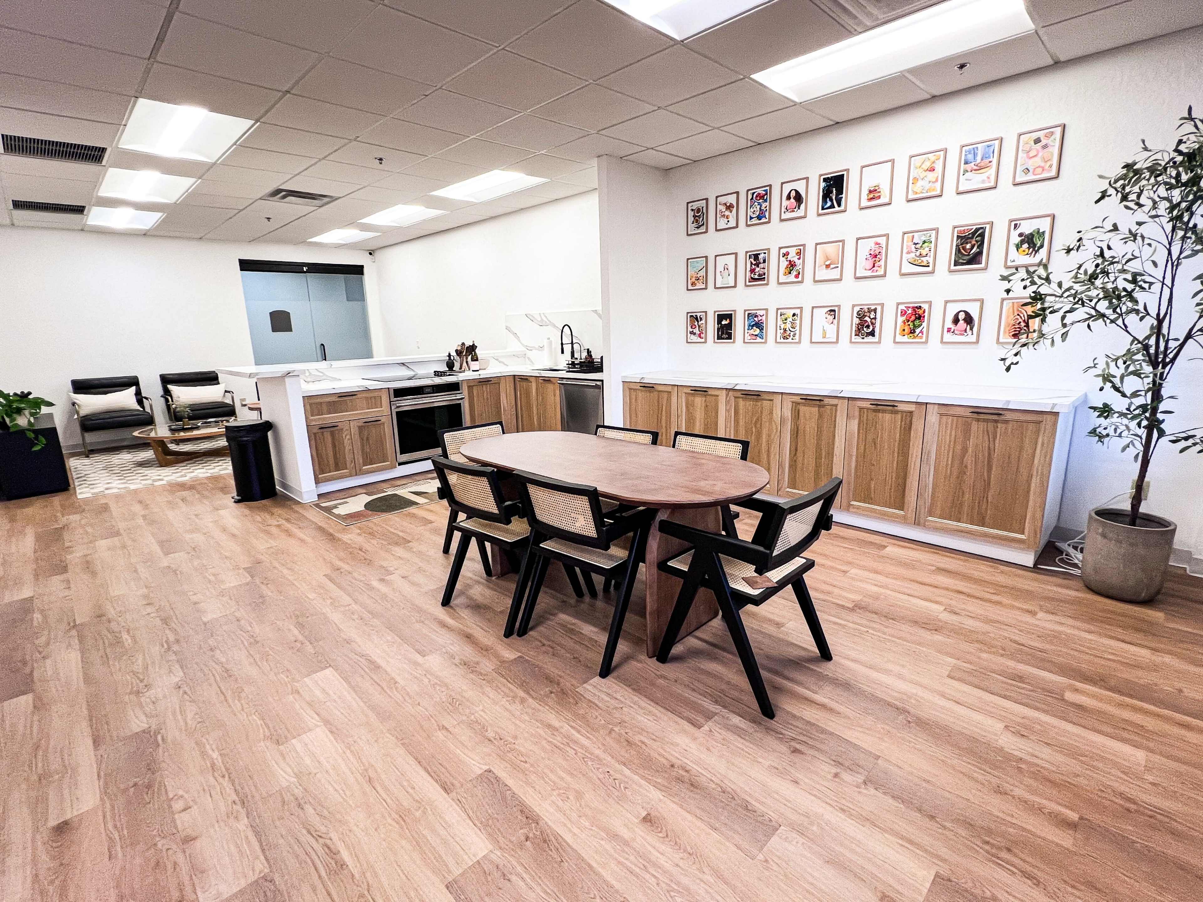 A modern office kitchen area features a wooden dining table surrounded by black chairs, with an art-adorned wall and a kitchen counter in the background.