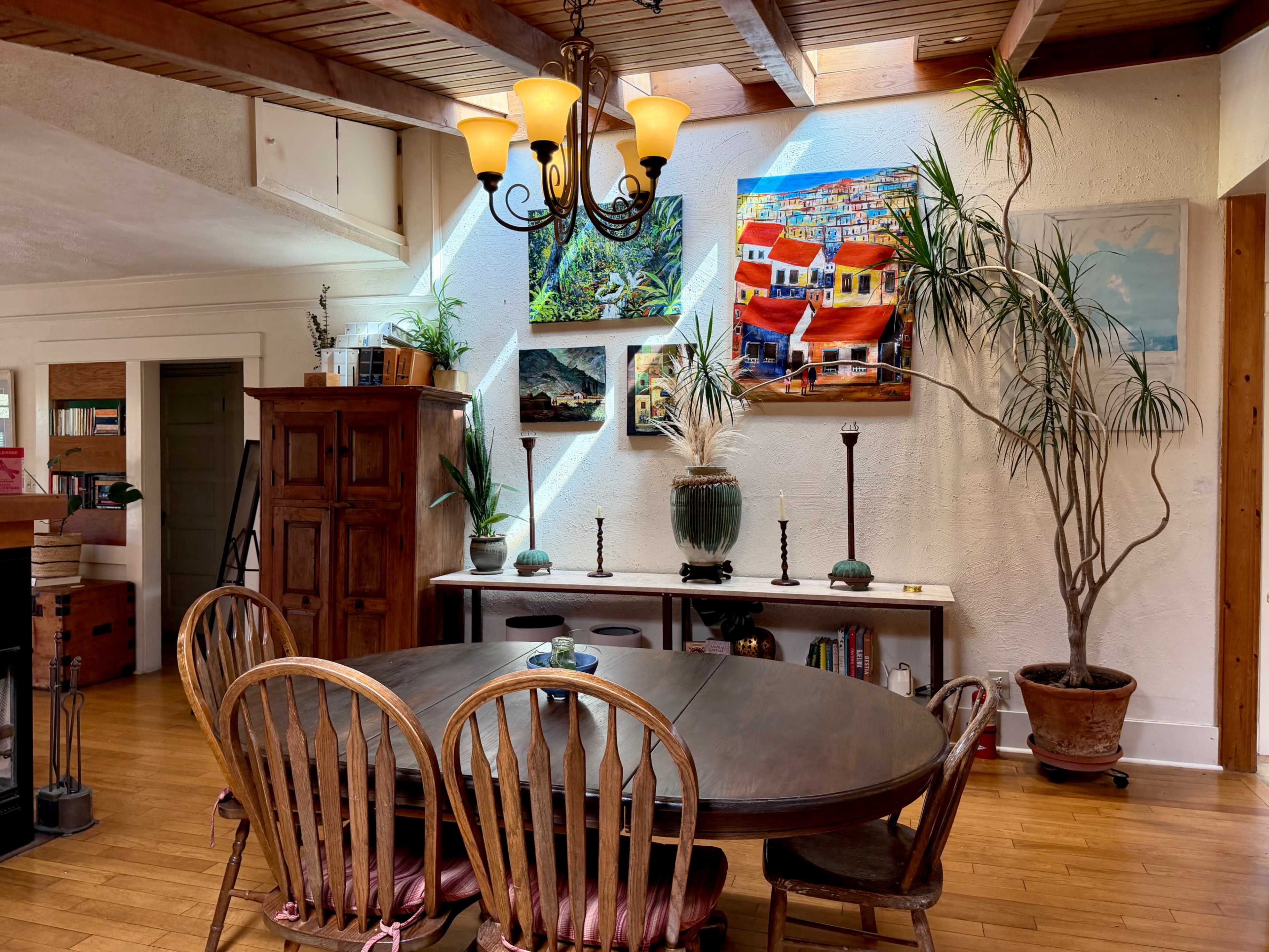 A dining area features a round wooden table surrounded by chairs, with colorful artwork mounted on the walls and potted plants adding greenery to the space.