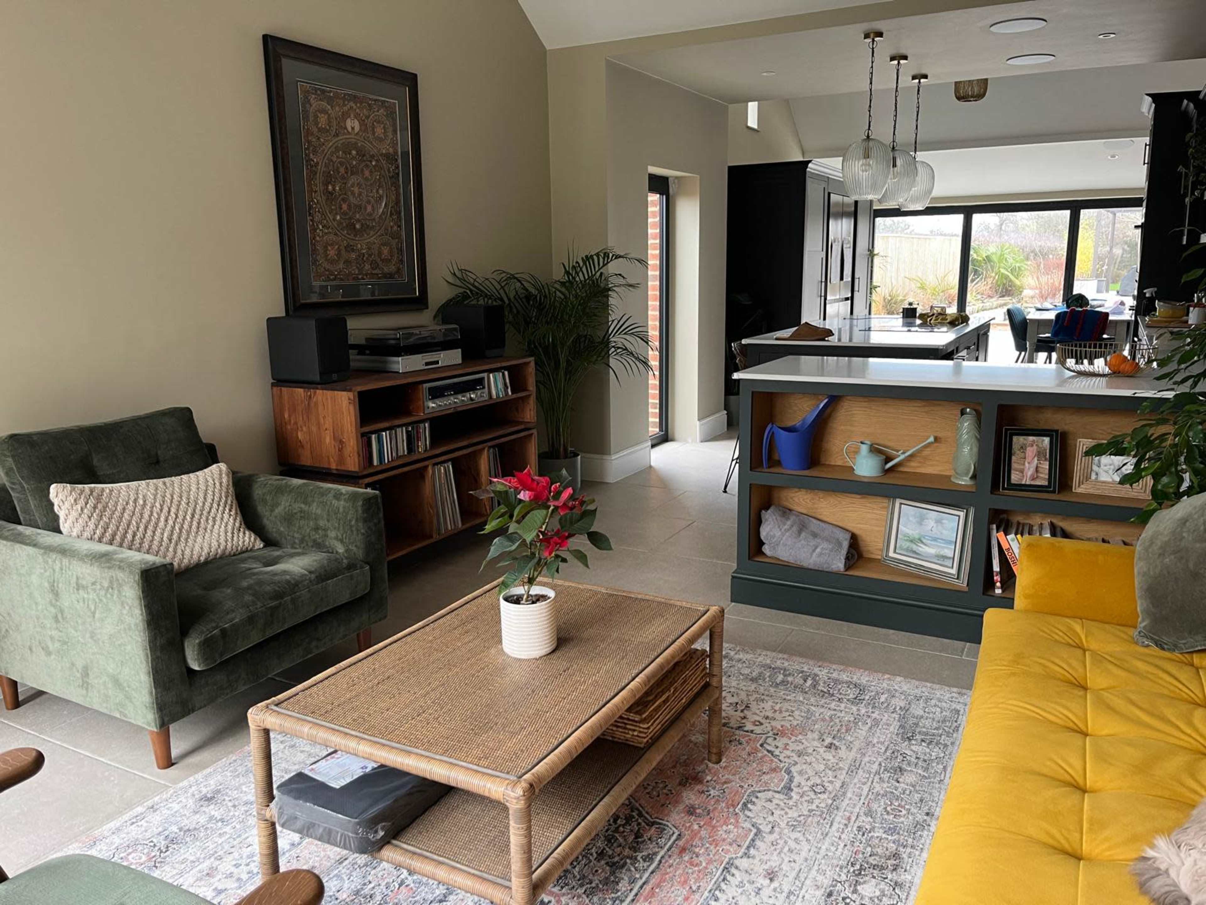 The image shows a modern living room with a green armchair, a yellow sofa, a wooden coffee table, and a view of a kitchen area in the background.