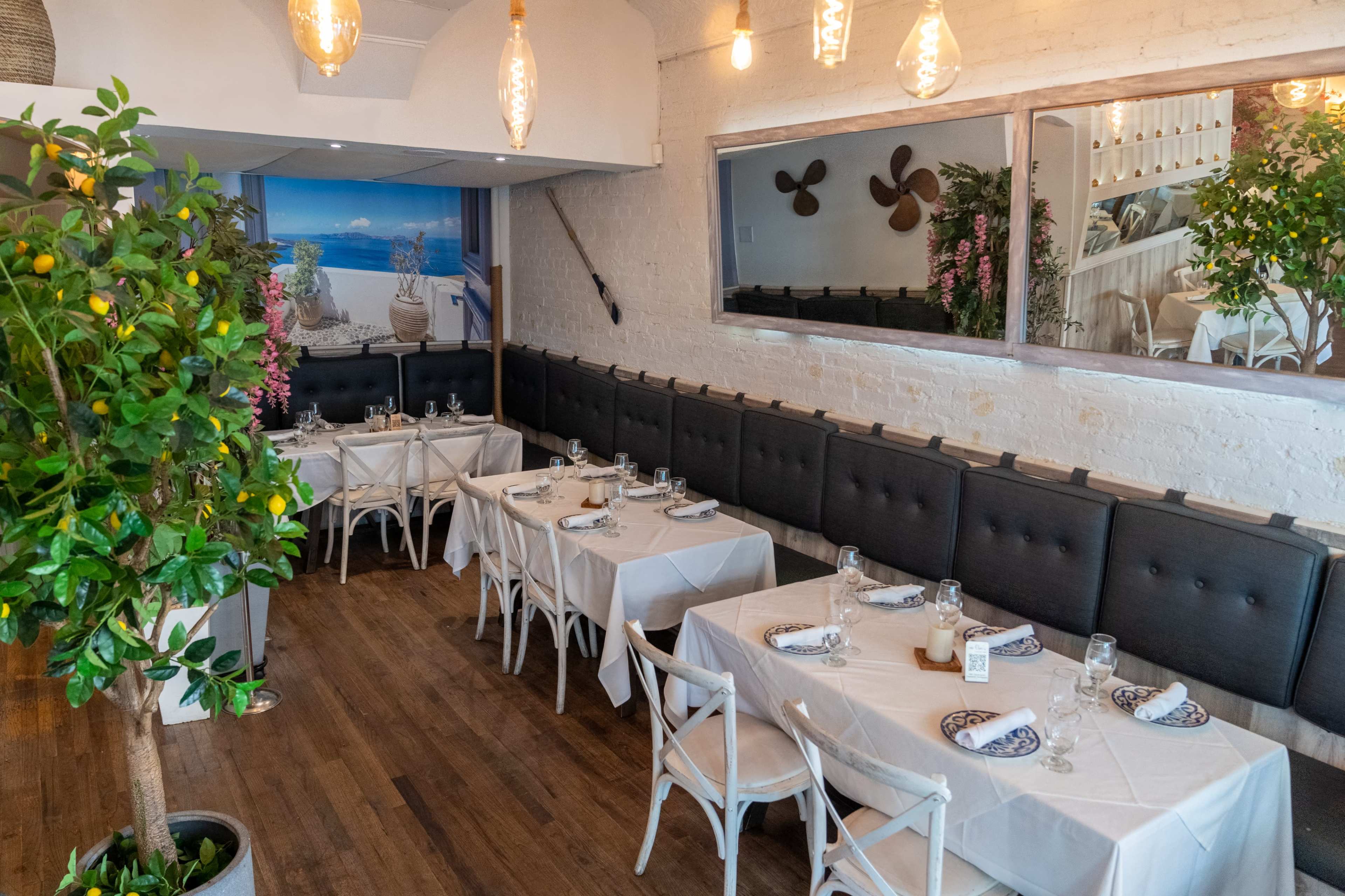 The image shows a bright restaurant interior with neatly arranged tables covered in white tablecloths, surrounded by greenery and decorative wall features.