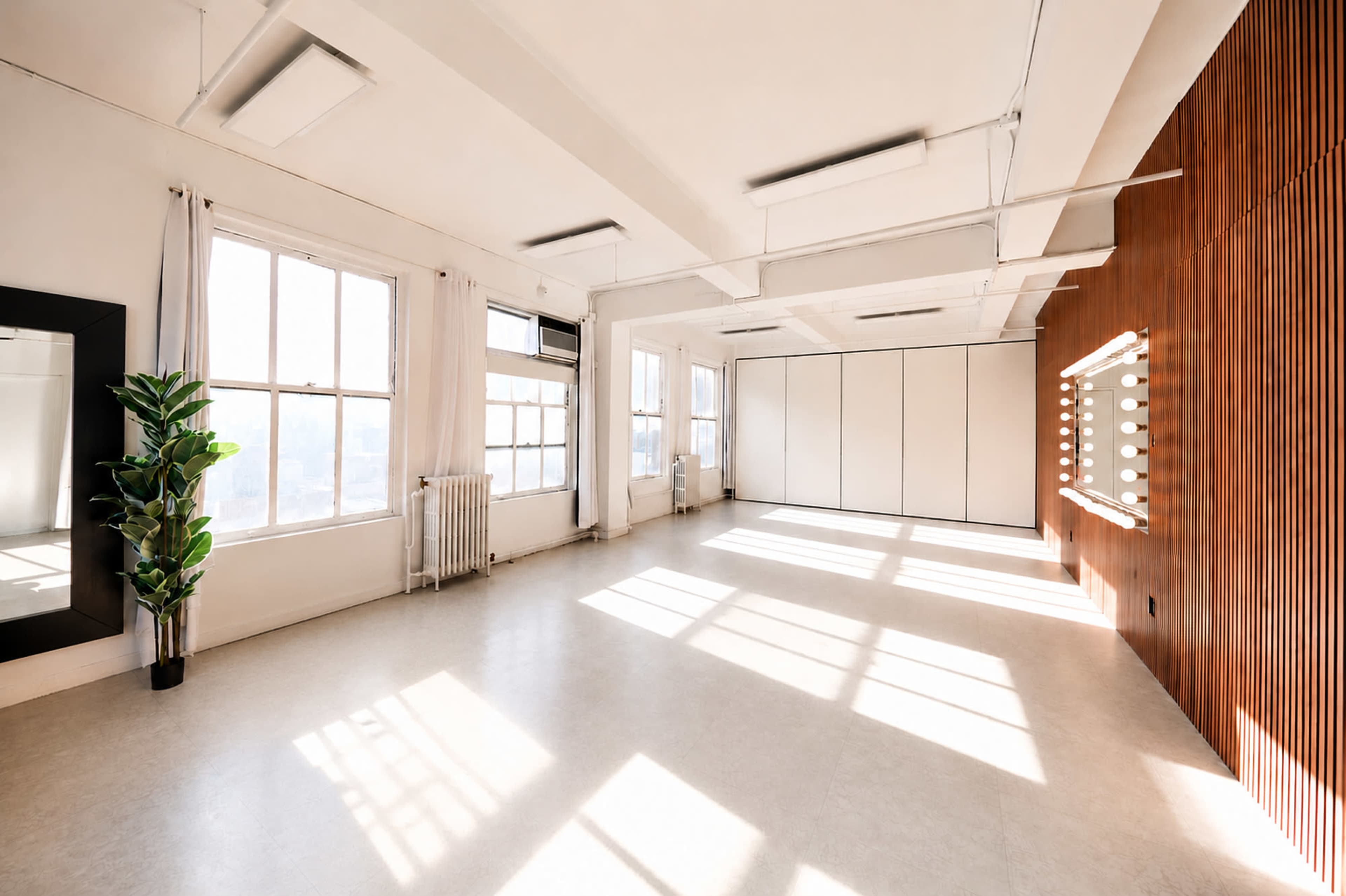 A bright, spacious room featuring large windows, a mirror, and a wall with light bulbs arranged around it.