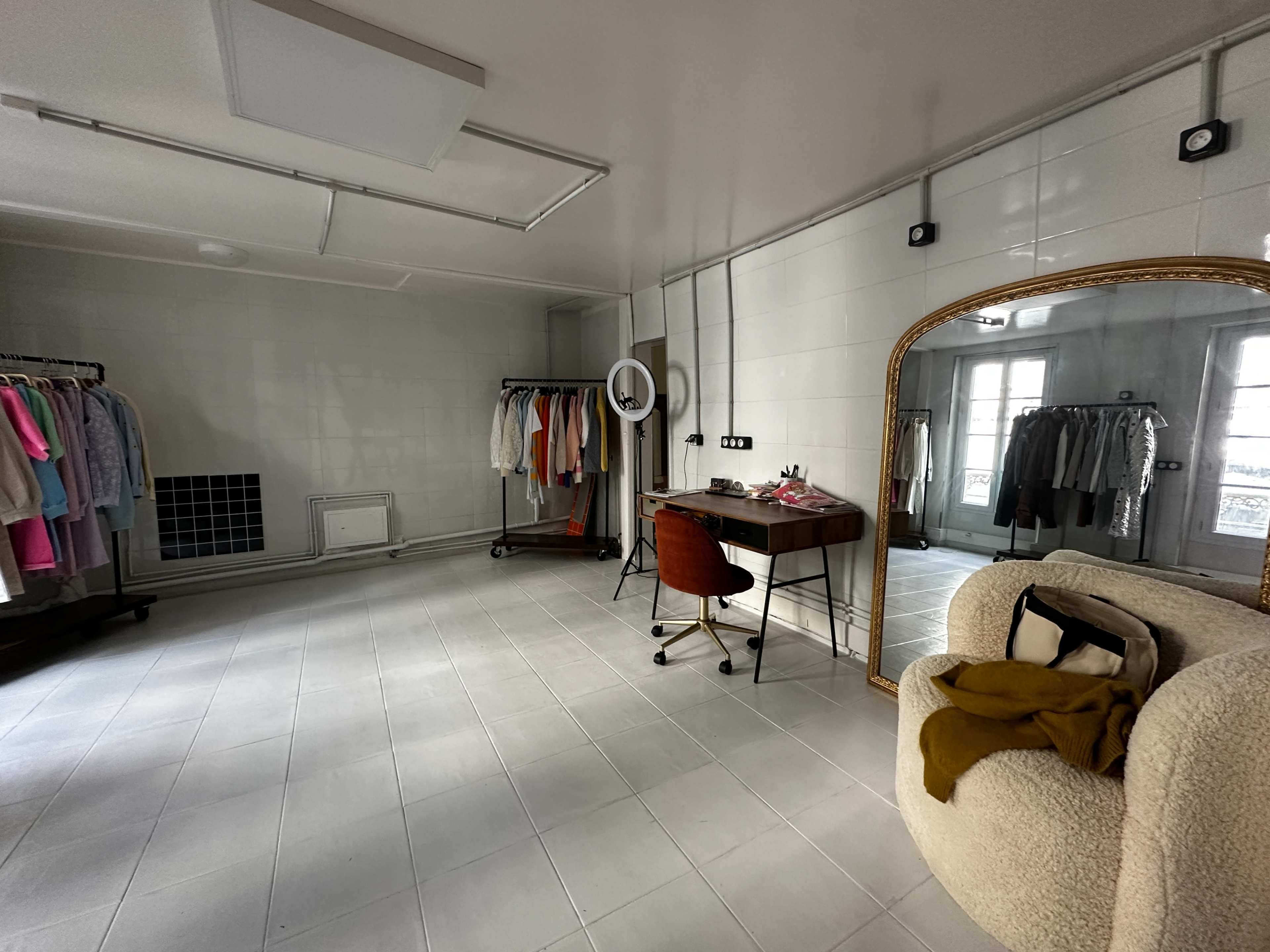 The image shows a spacious, minimalist room with clothing racks on one side, a desk with a chair in the center, and a large mirror on the opposite wall.