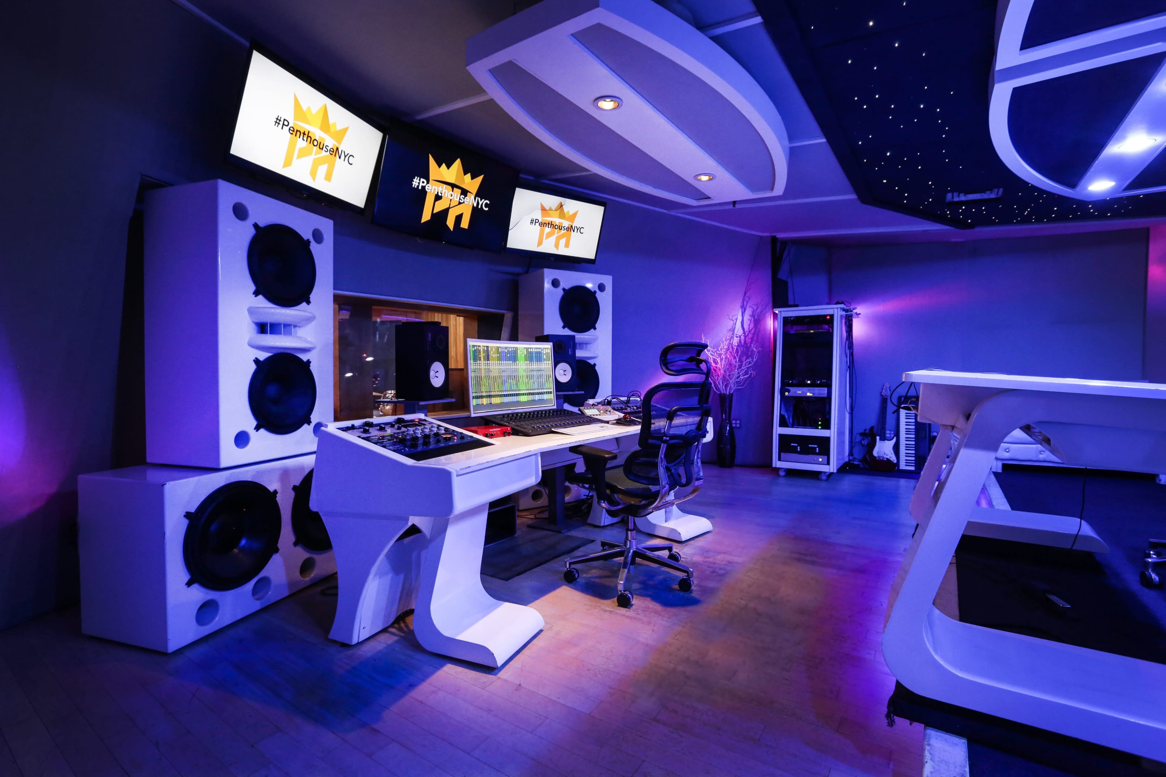 The image shows a modern music studio featuring large speakers, a digital mixing console, and multiple screens displaying audio software, all illuminated by purple lighting.