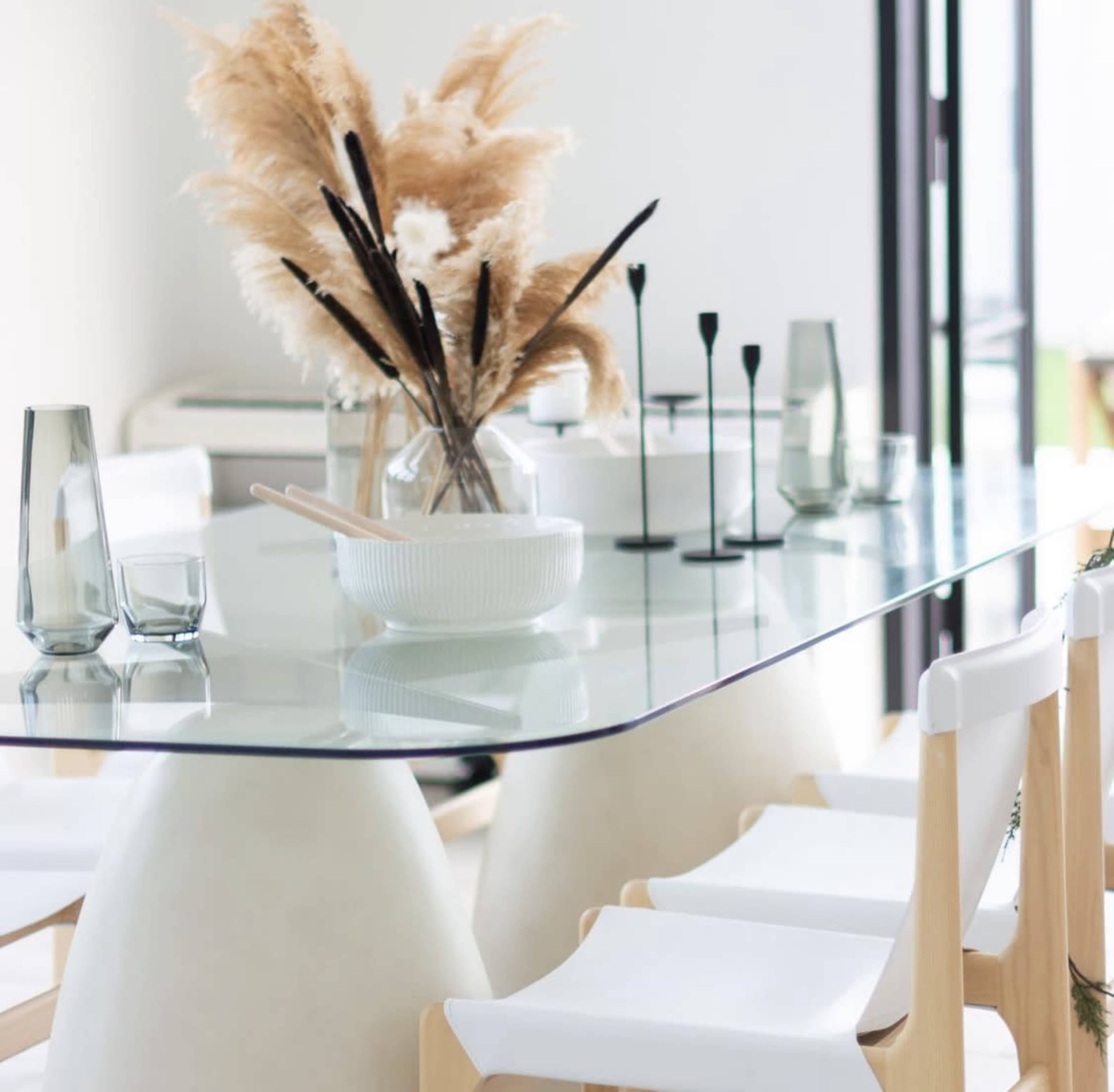 A glass dining table is set with minimalist decor, including a vase of dried pampas grass and various glassware, surrounded by white chairs.