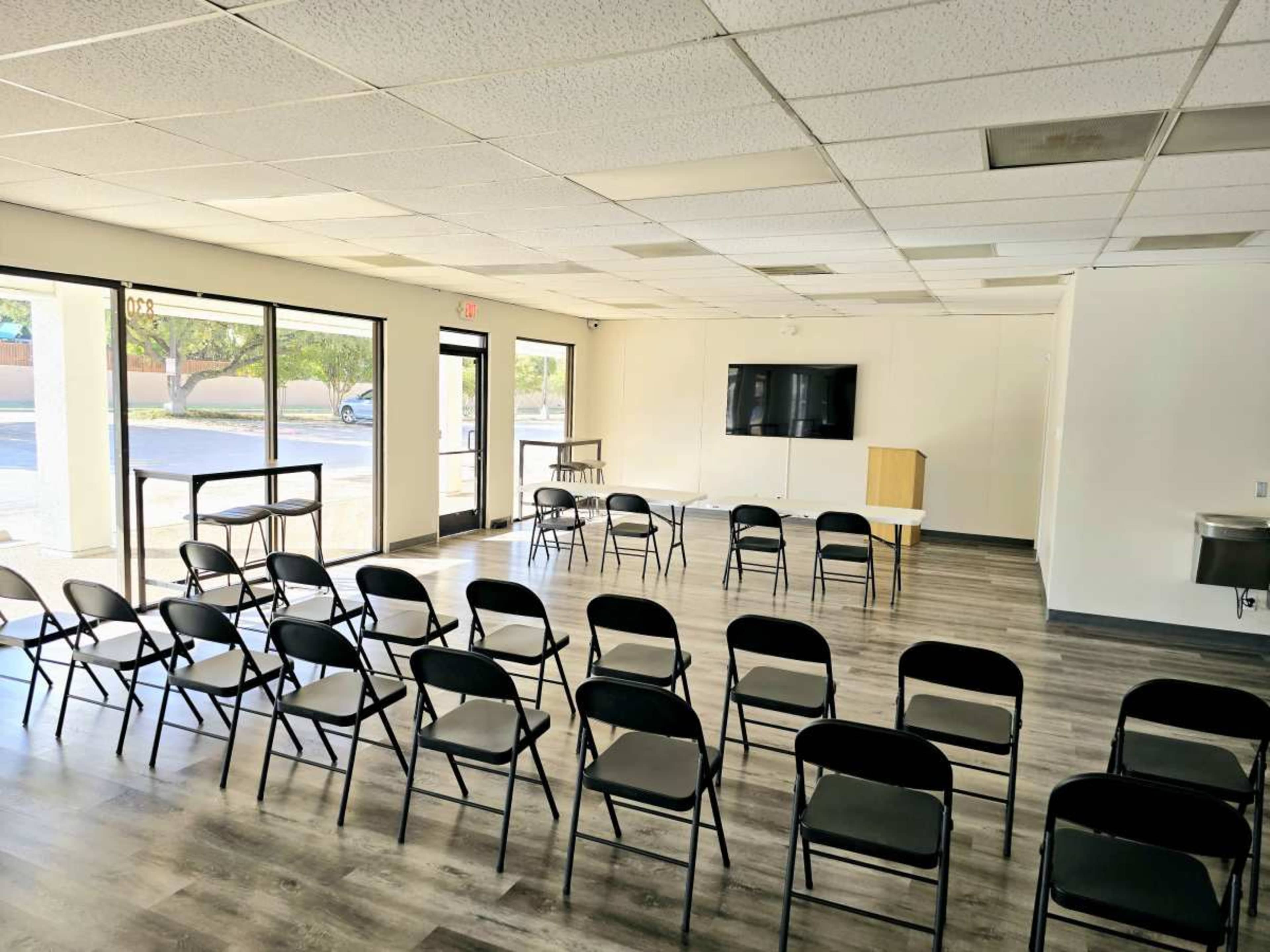 The image shows a meeting room with rows of black chairs facing a table and a television mounted on the wall.