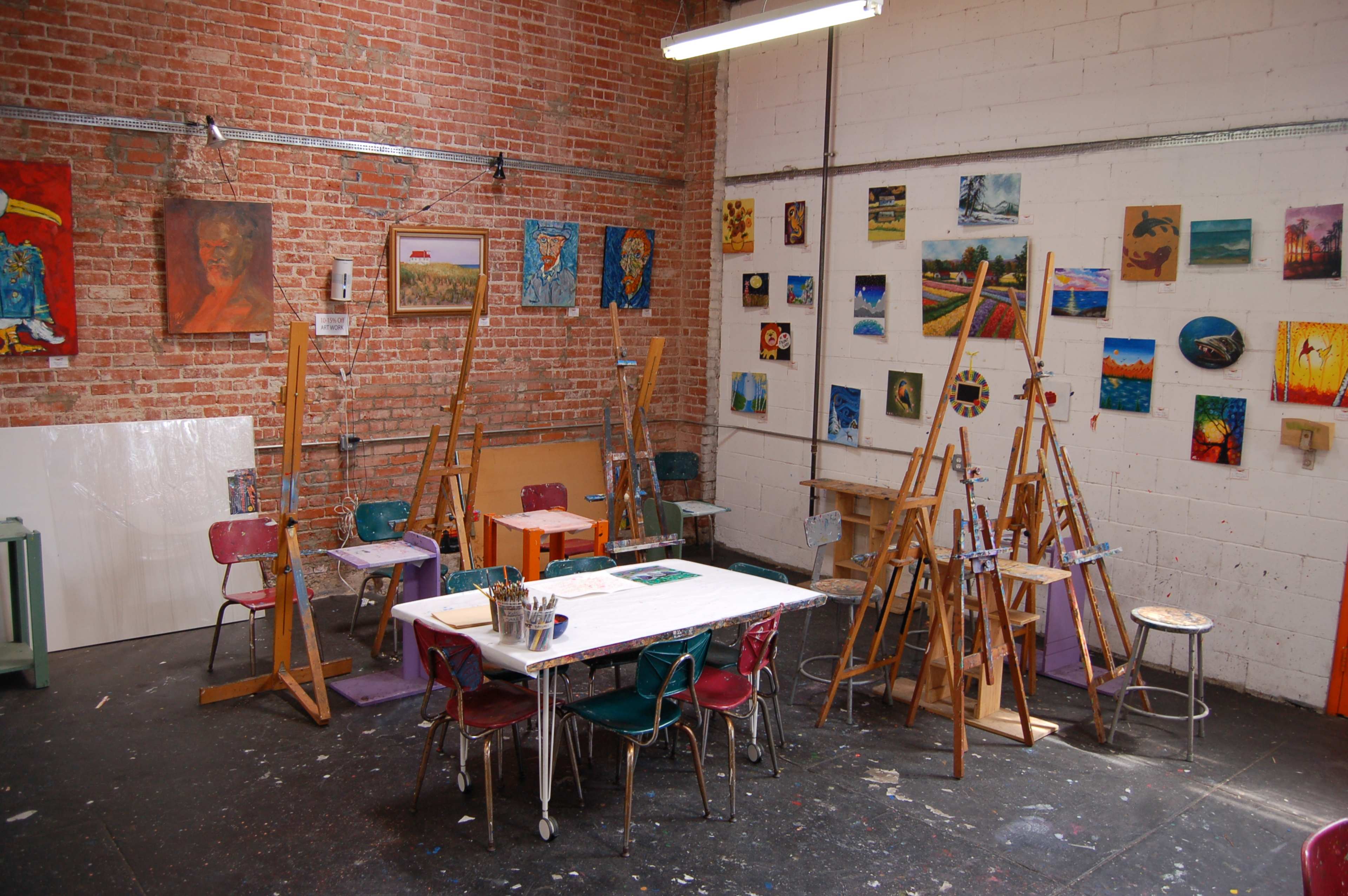 An artist studio filled with easels, chairs, tables, and various paintings displayed on the brick walls.