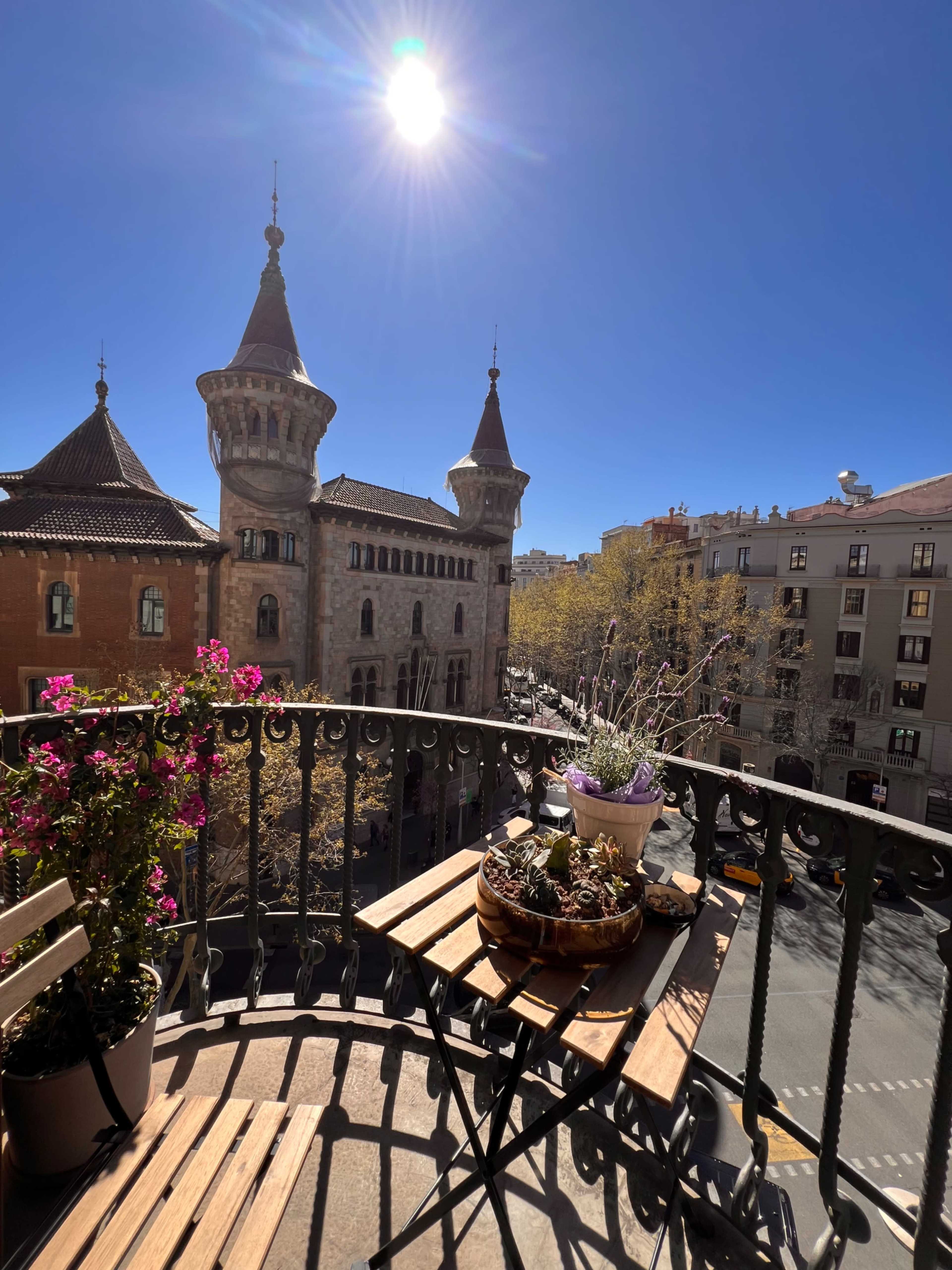 A sunny balcony features potted plants and wooden furniture, overlooking a street with historic buildings.
