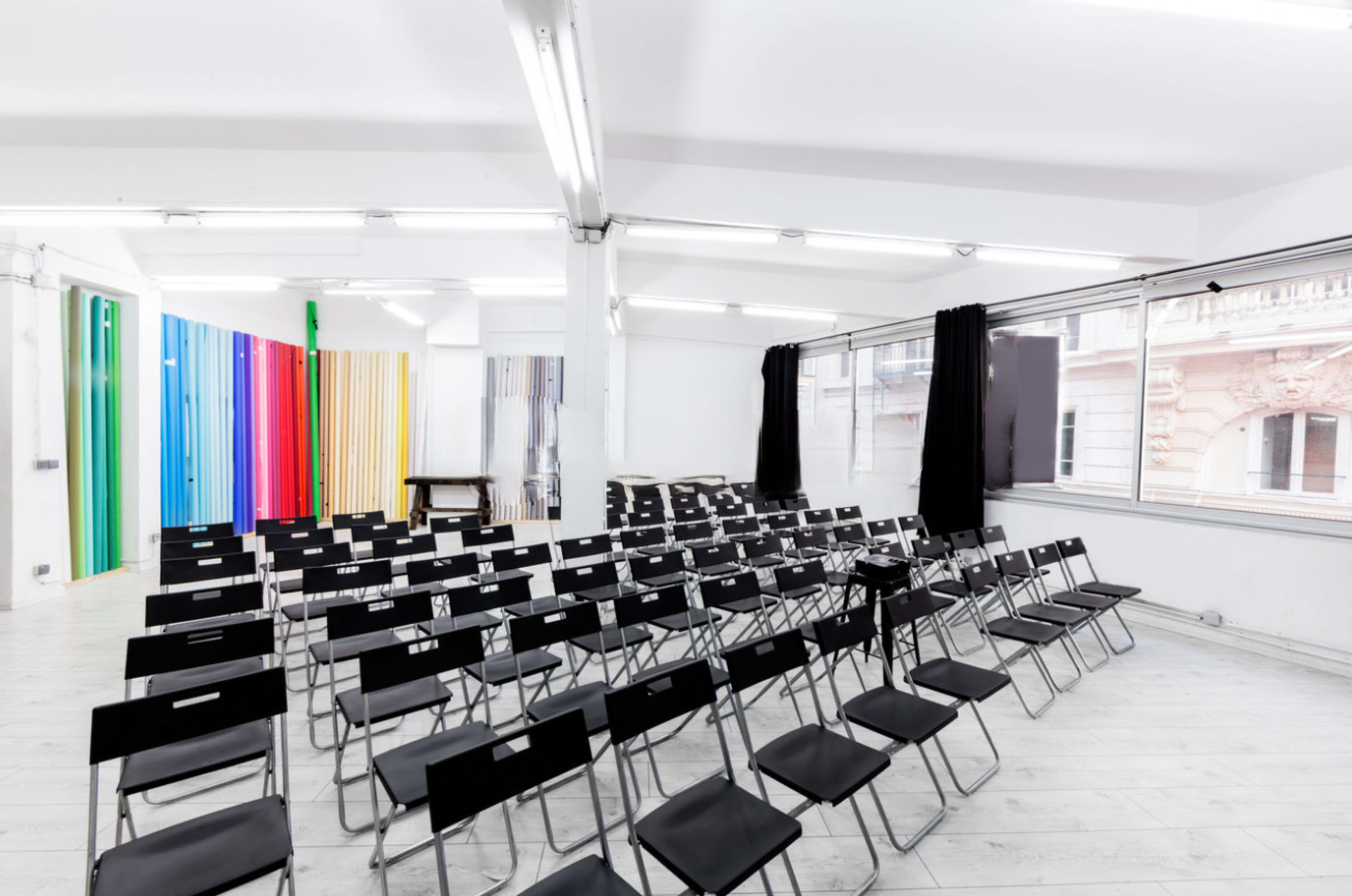 An empty room filled with rows of black folding chairs facing a blank wall, with colorful panels in the background.