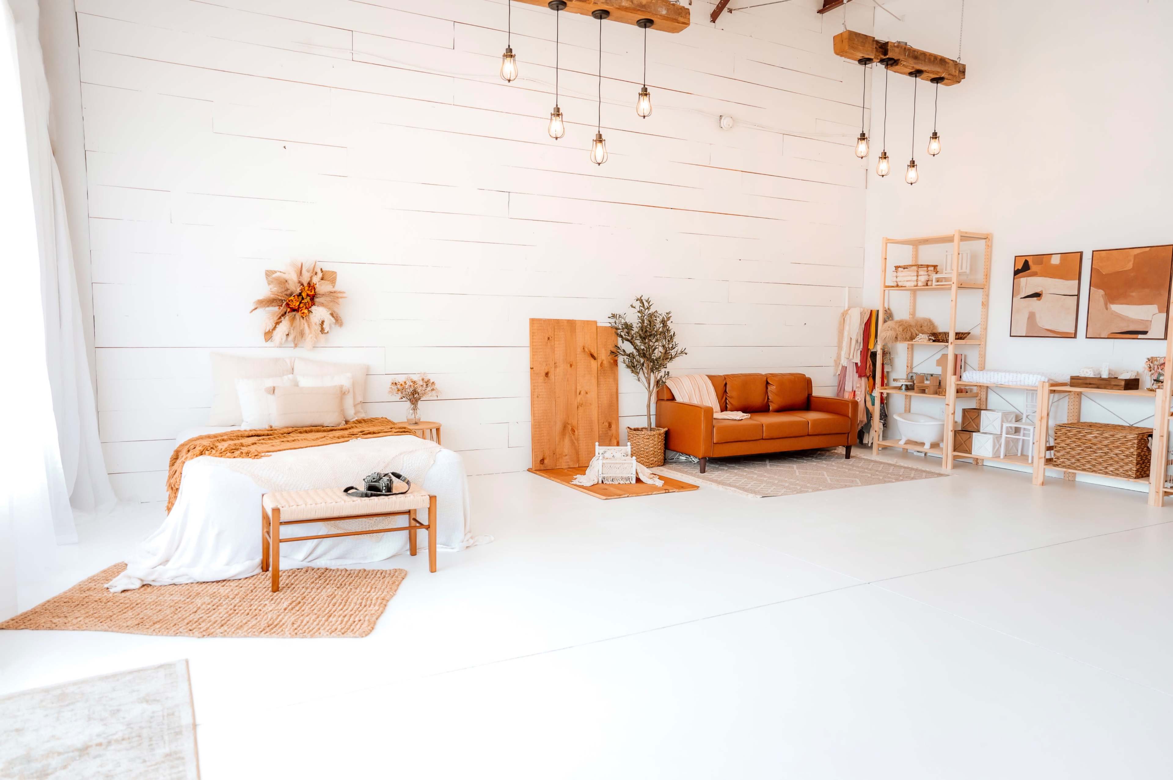 The image depicts a bright, minimalistic interior space featuring a bed with white bedding, a brown sofa, wooden furniture, and decorative plants.