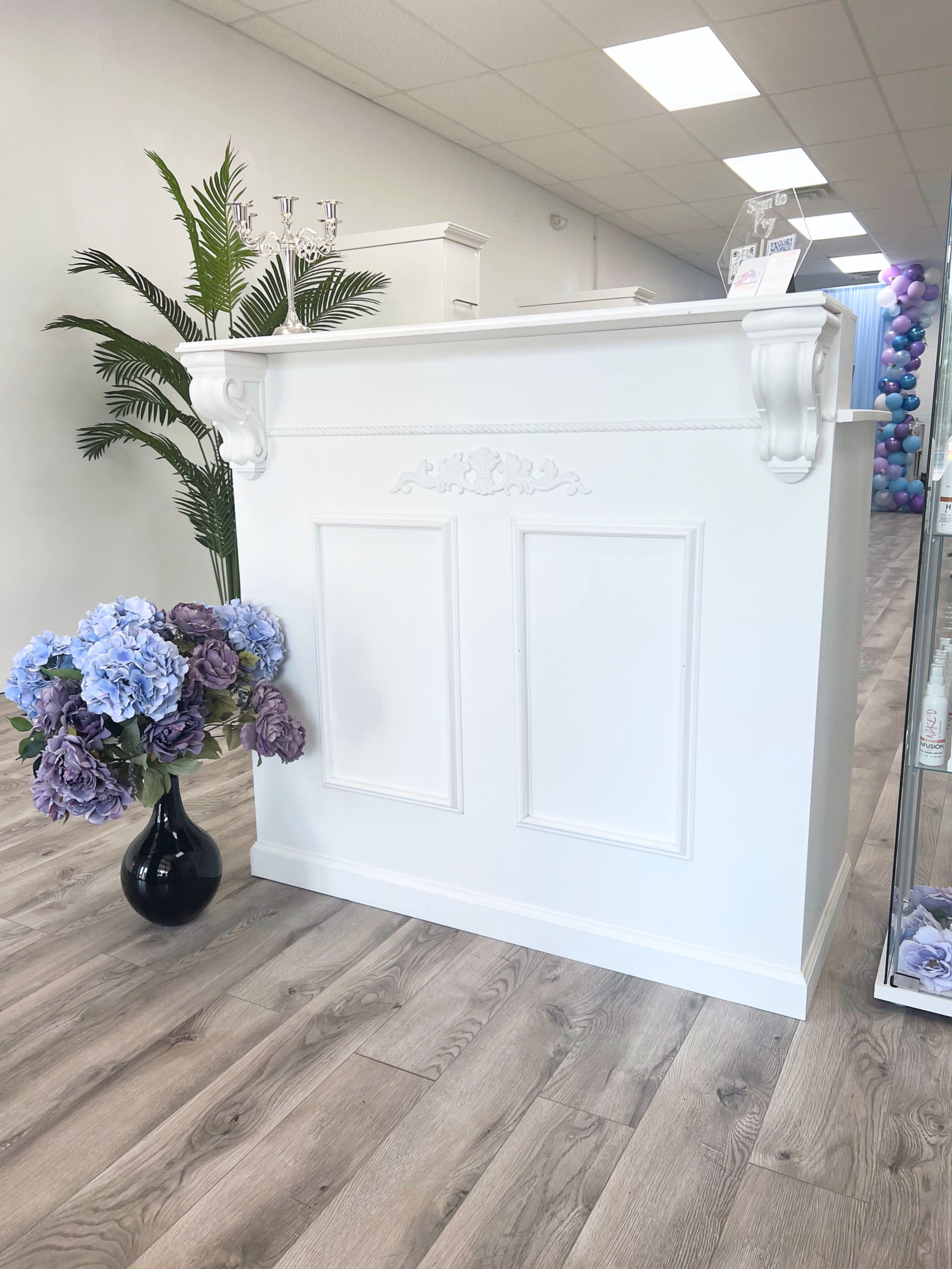 A white reception desk with ornate features, accompanied by a black vase filled with blue and purple flowers, set in a bright, modern interior.