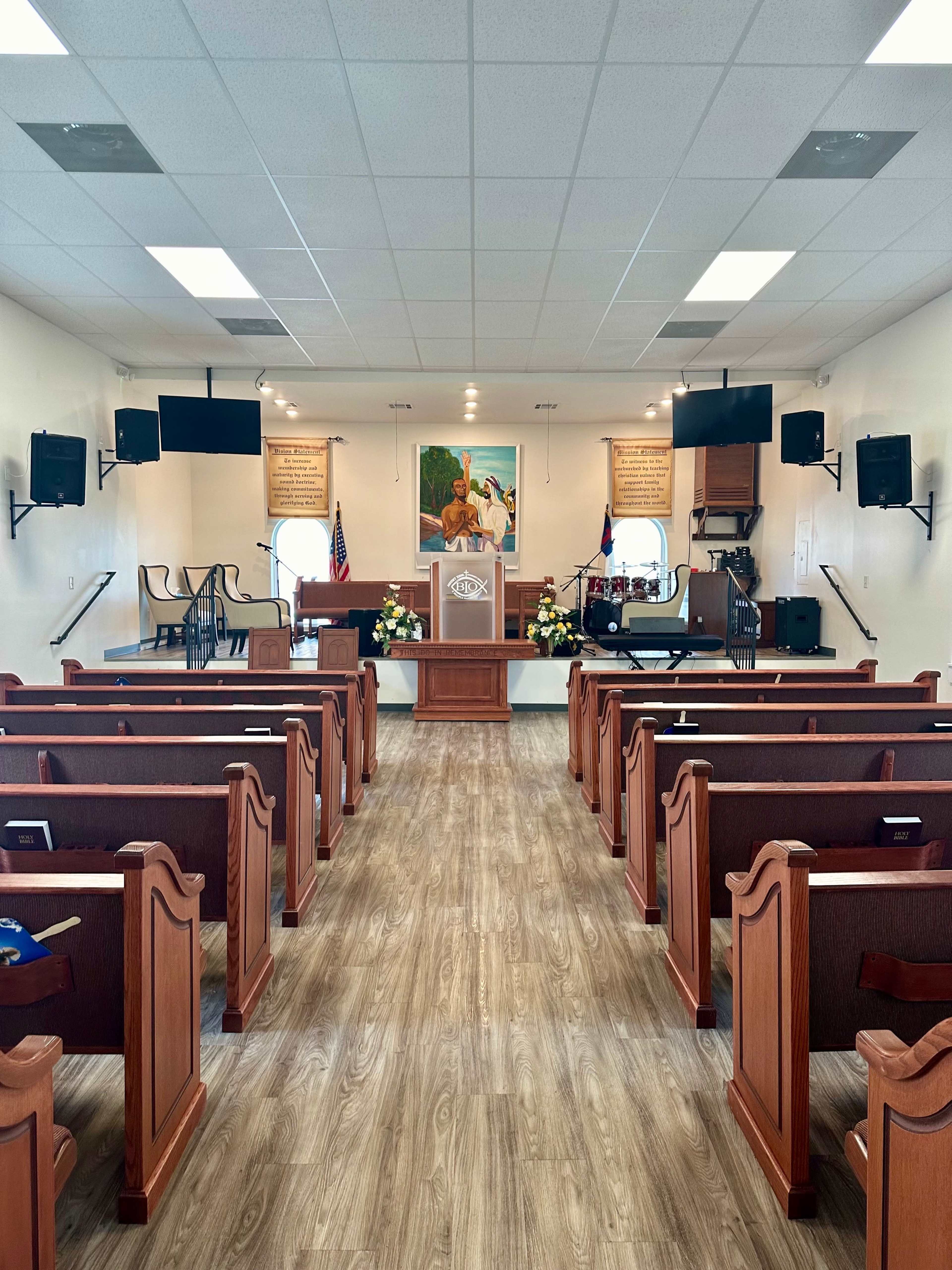 The interior of a church features wooden pews facing a podium, adorned with floral arrangements and wall decorations.