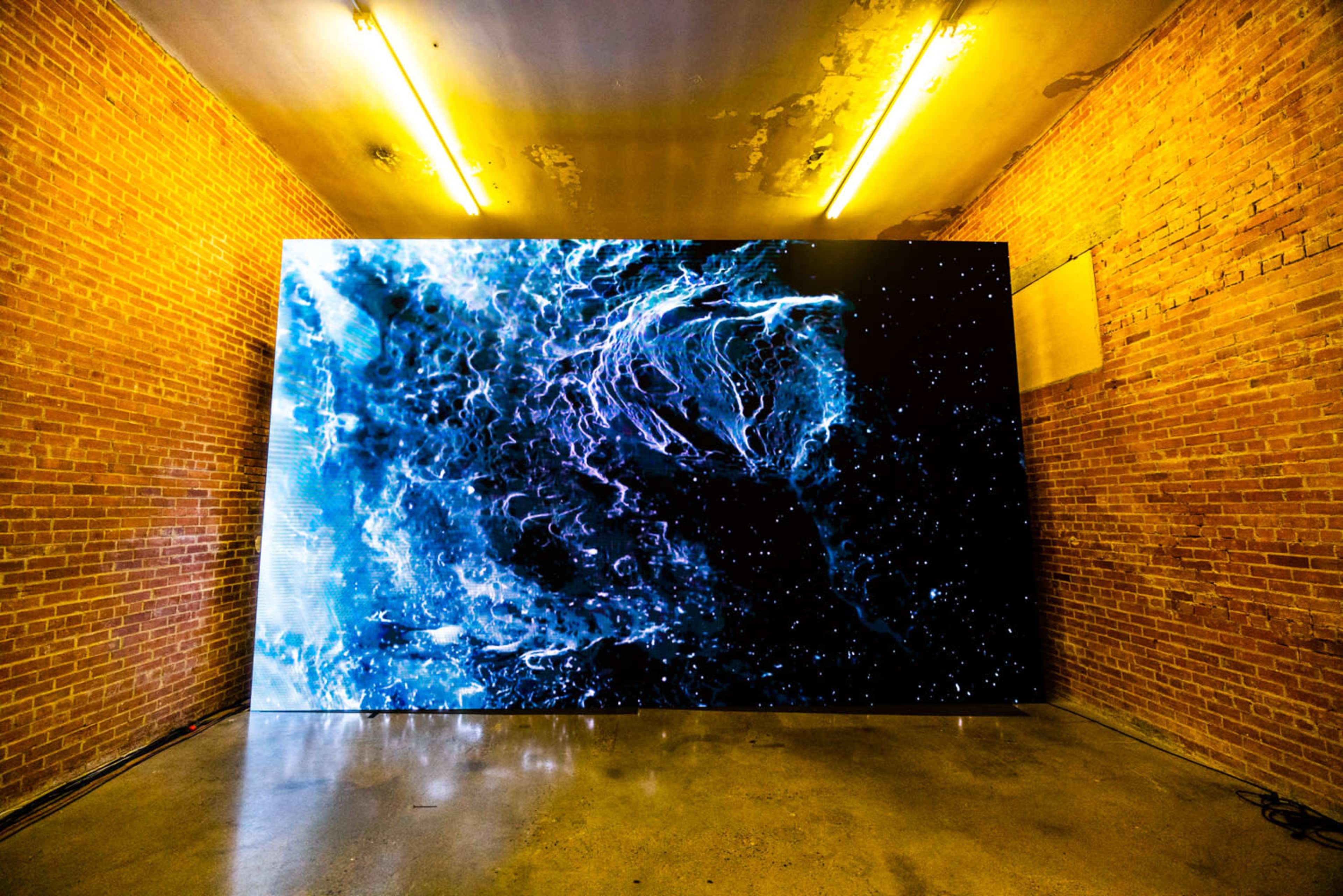 A large digital screen displaying a vibrant abstract visual is installed on a brick wall in a dimly lit room with exposed lighting fixtures.