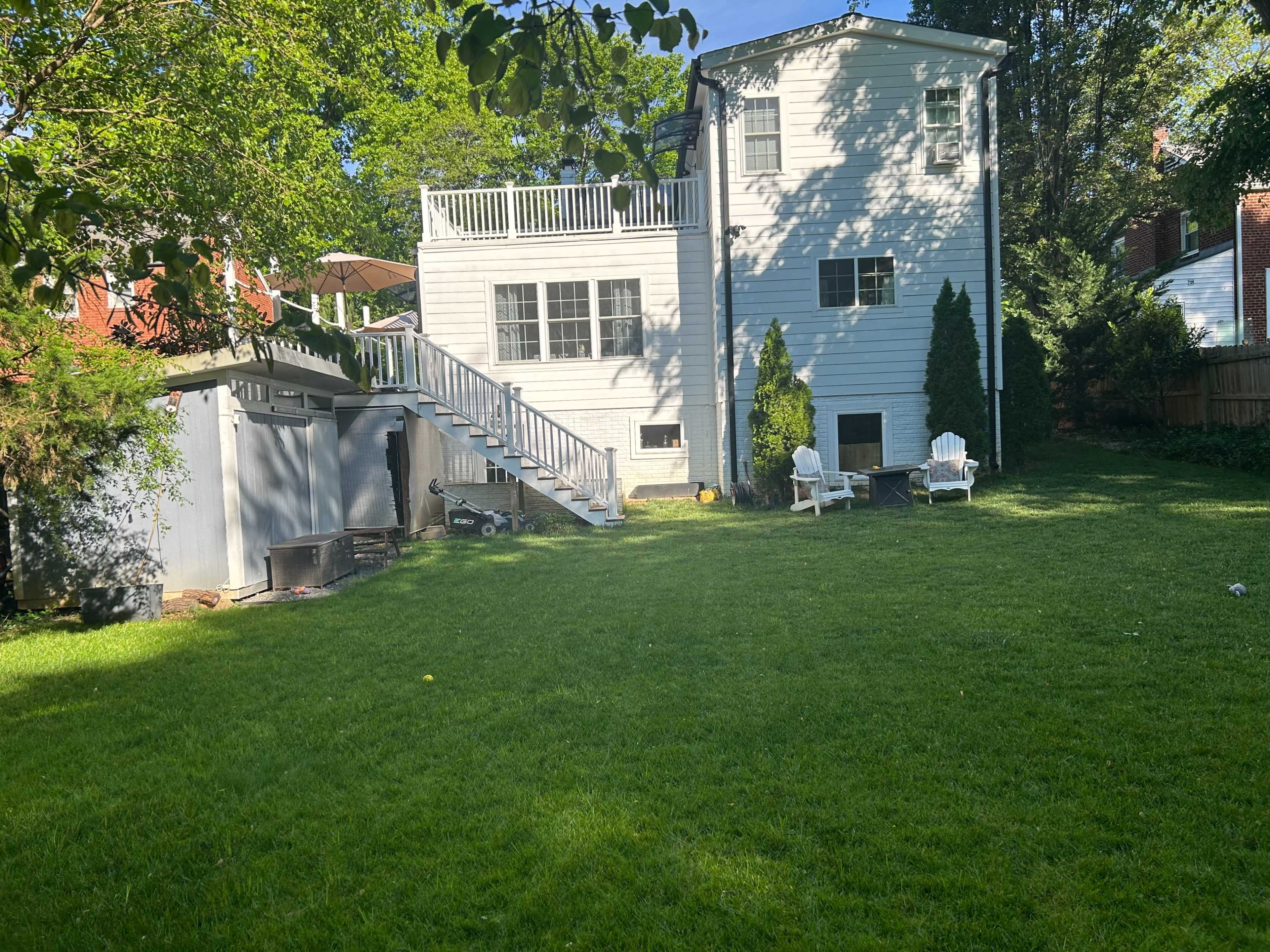A two-story house with white siding and a deck overlooks a green lawn, which is bordered by trees and features outdoor seating.