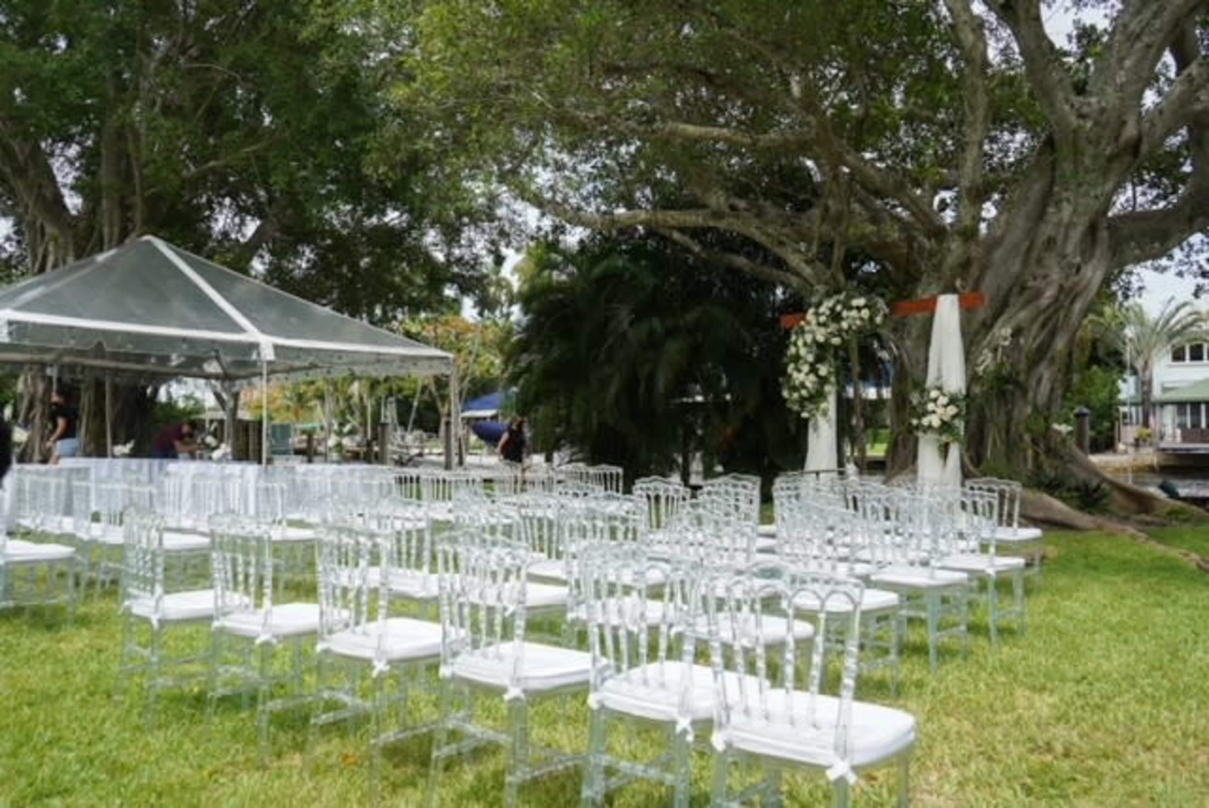 A wedding setup with transparent chairs arranged in rows beneath a tent, next to a decorated archway and large trees.