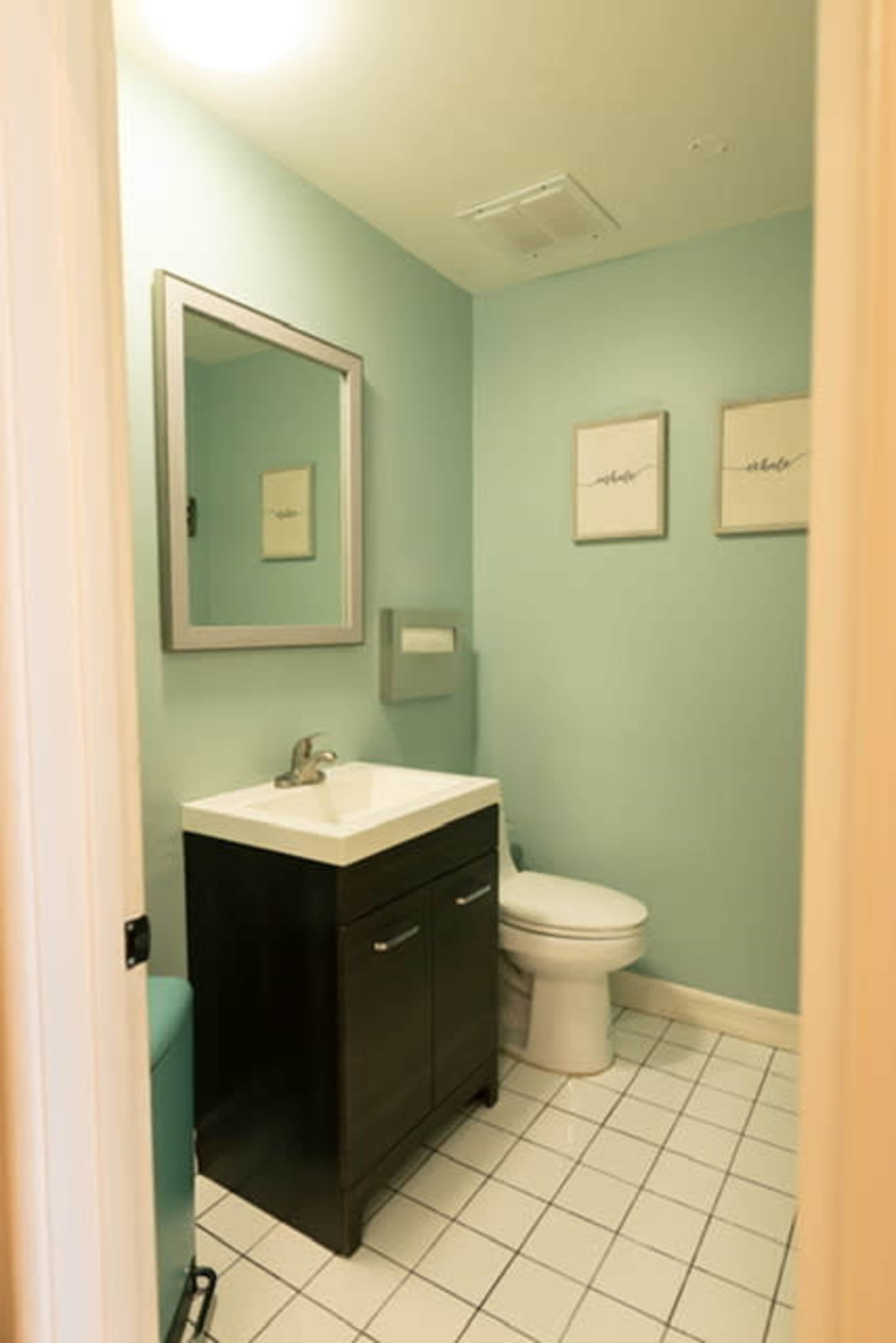 The image shows a compact bathroom featuring a white vanity with a sink, a toilet, light blue walls, and two framed pictures on the wall.