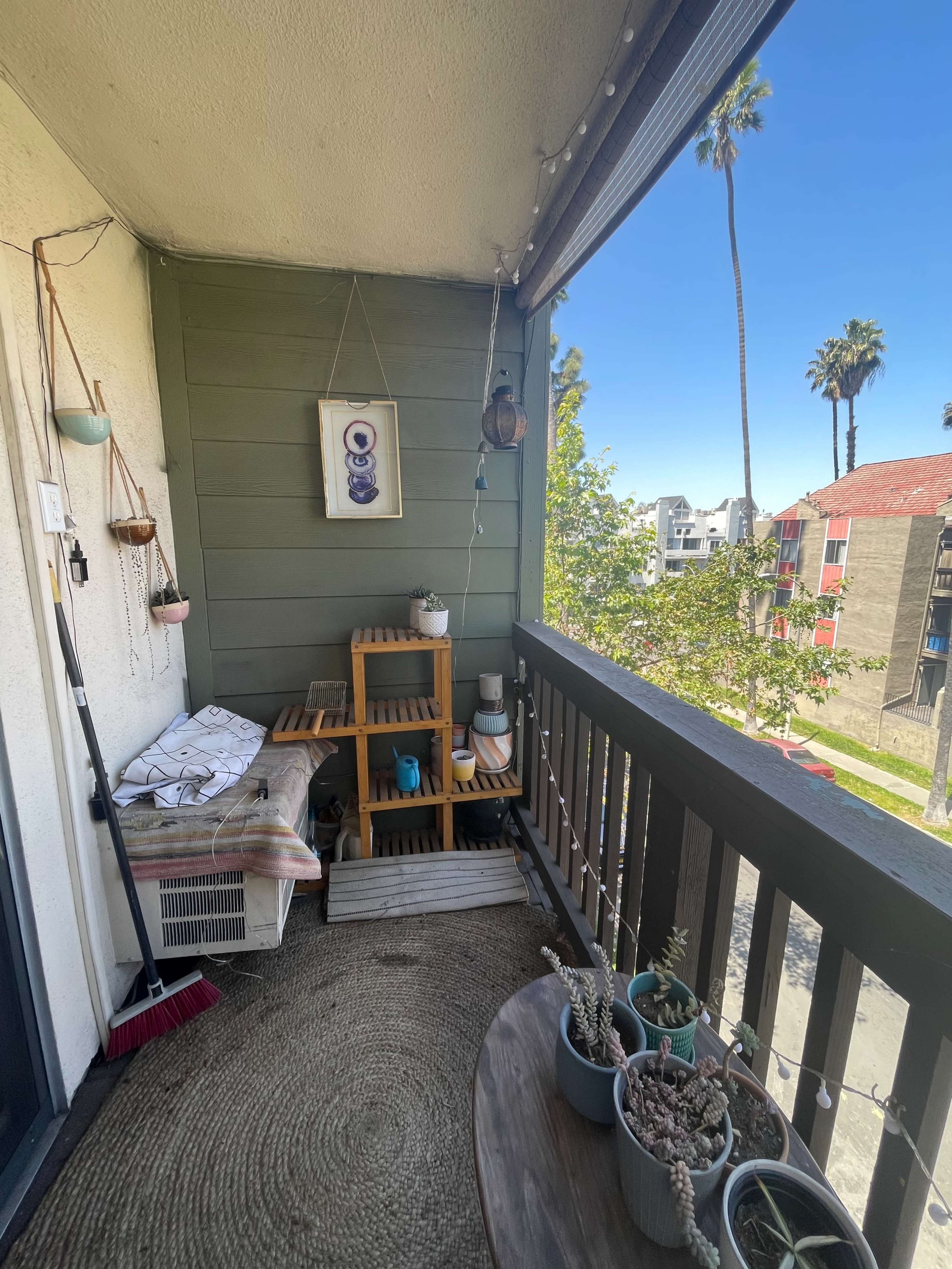 A small balcony features a wooden shelf with various plants, a seating area, and a view of palm trees and nearby buildings.