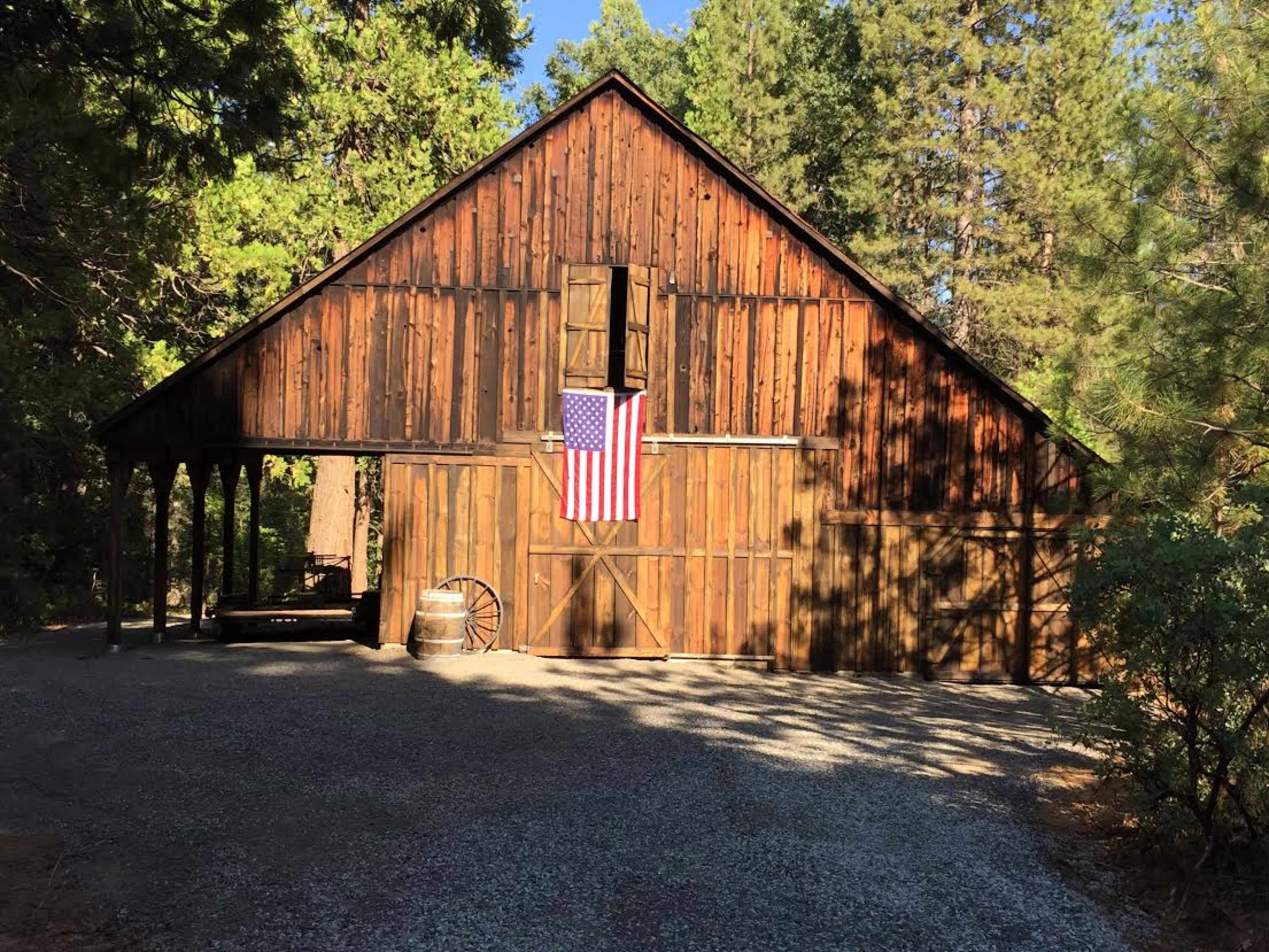 A rustic wooden barn with an American flag hanging on its front, set in a wooded area.