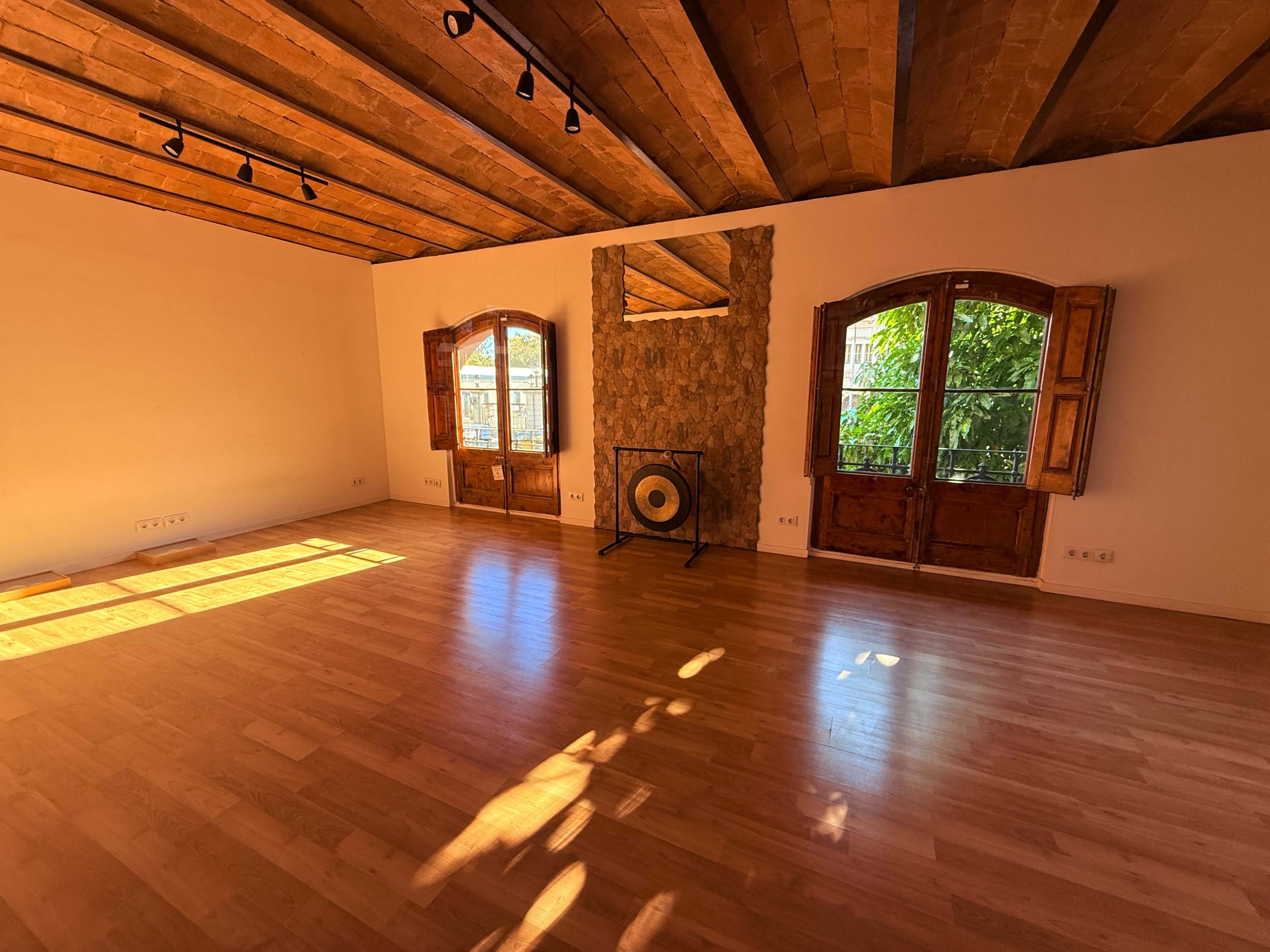 The room features wooden beams on the ceiling, a stone wall with a mirror, a large gong, and two open windows allowing natural light to enter.