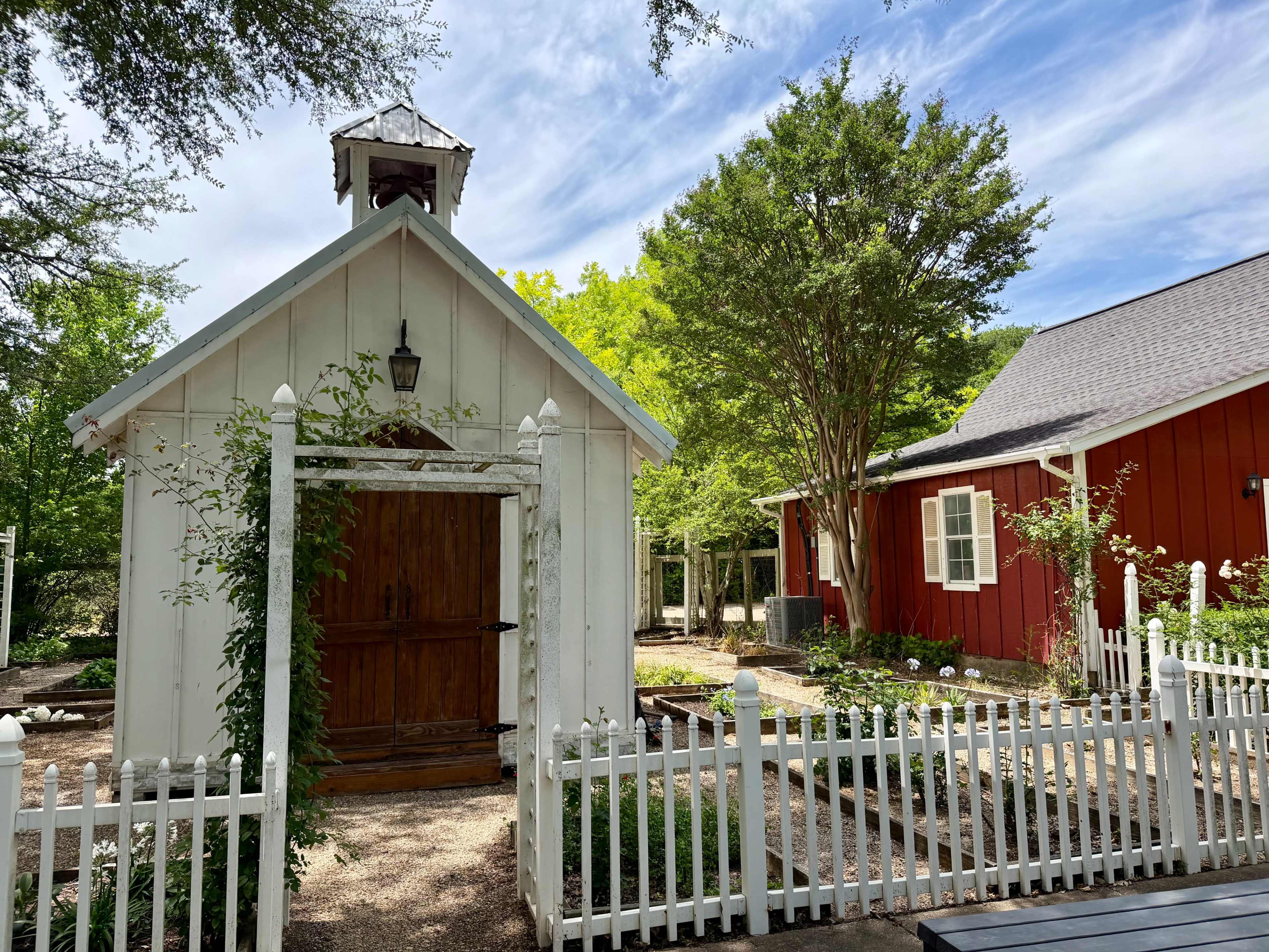 The image shows a white shed with a bell tower next to a red building in a landscaped garden with a white picket fence.