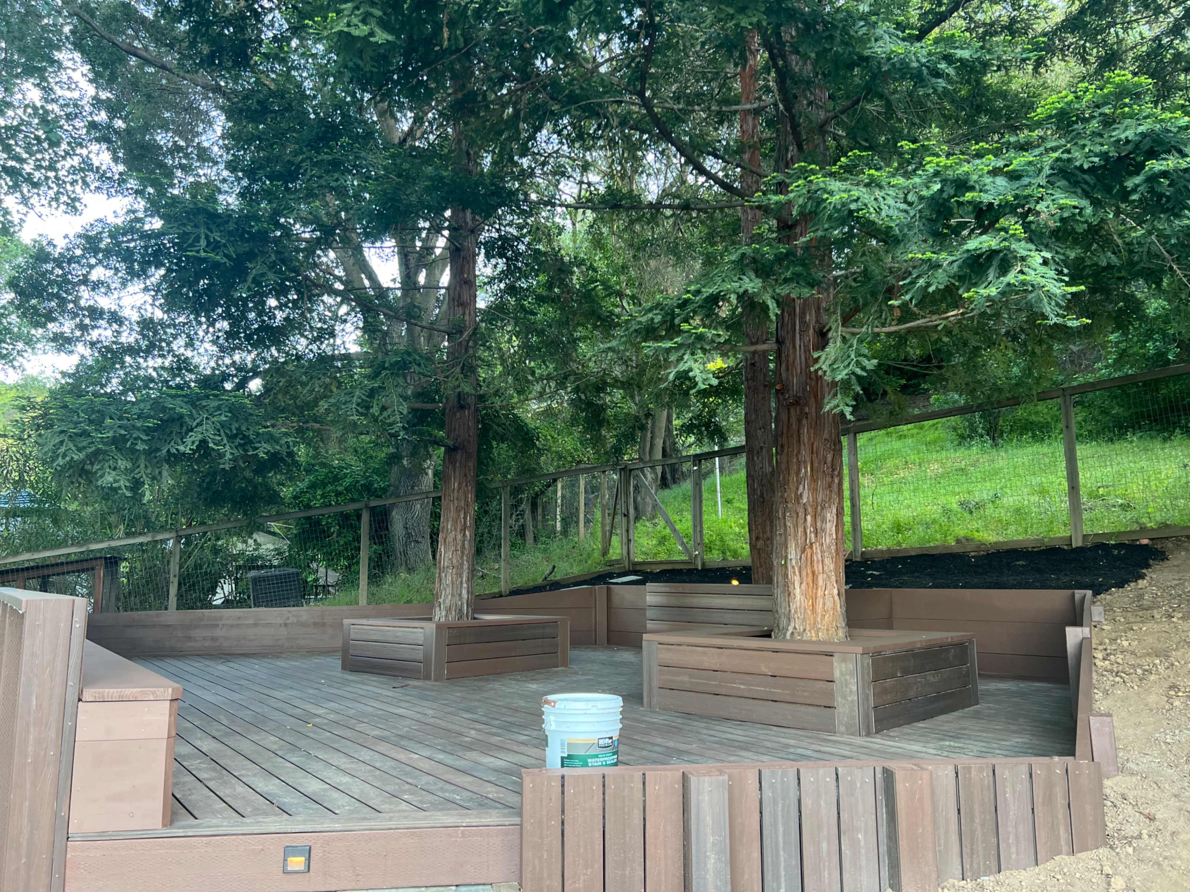 The image shows a wooden deck area surrounded by trees, featuring raised planter boxes and a bucket on the ground.