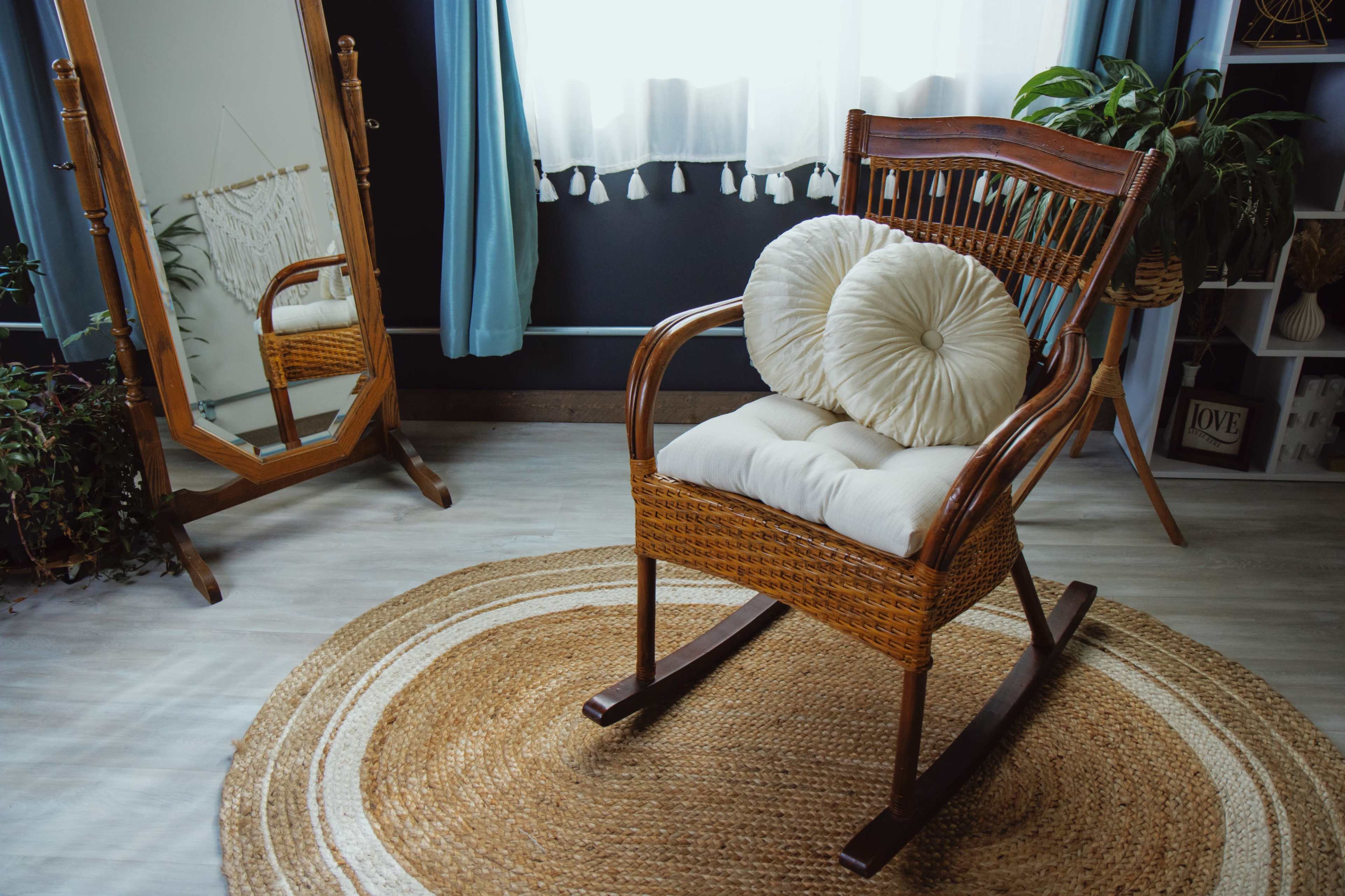 A wooden rocking chair with decorative cushions sits on a circular jute rug in a room with a large mirror and potted plants.