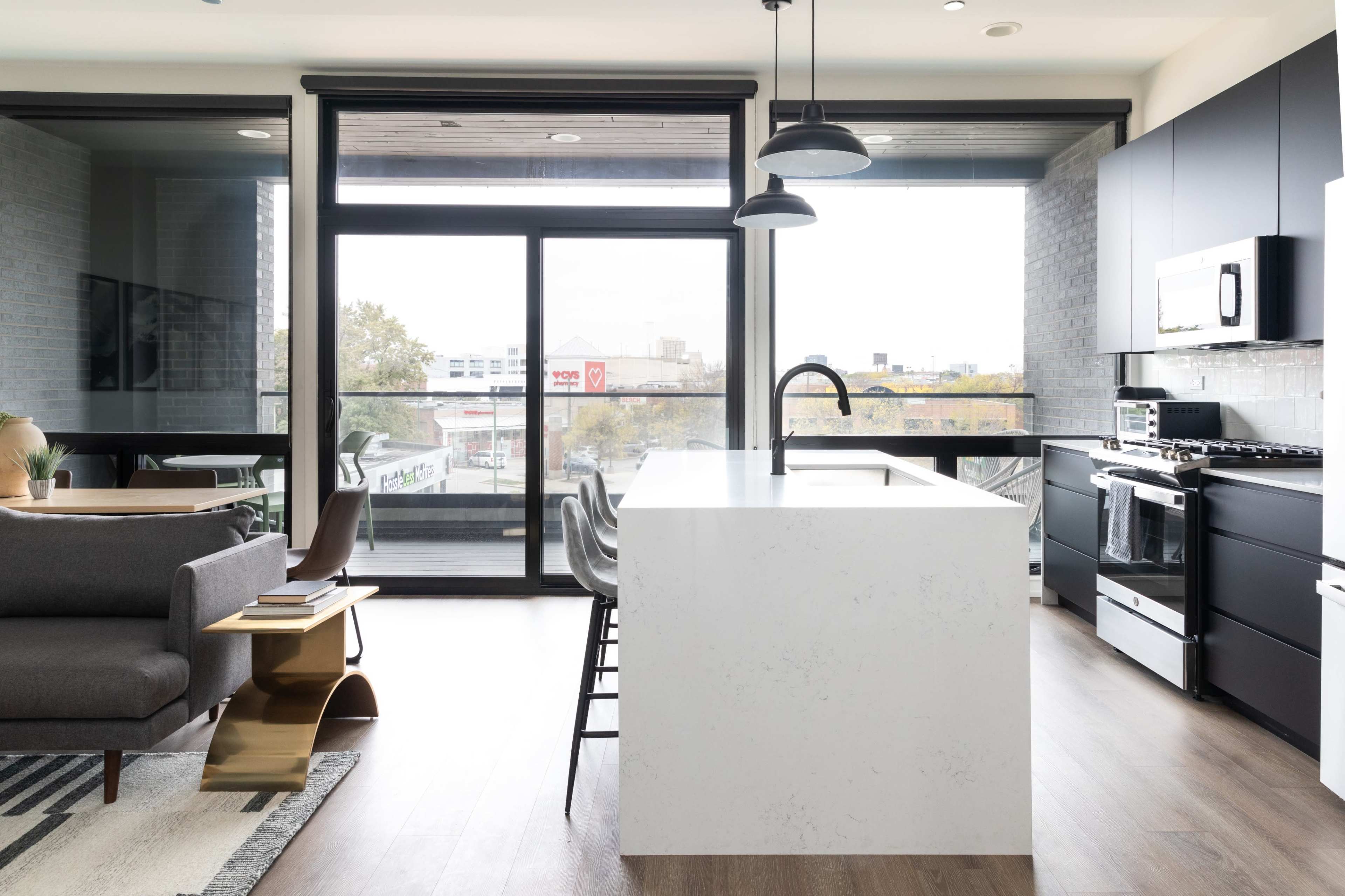 A modern kitchen features a large white island with a sink, surrounded by bar stools, and opens to a living area with large windows.