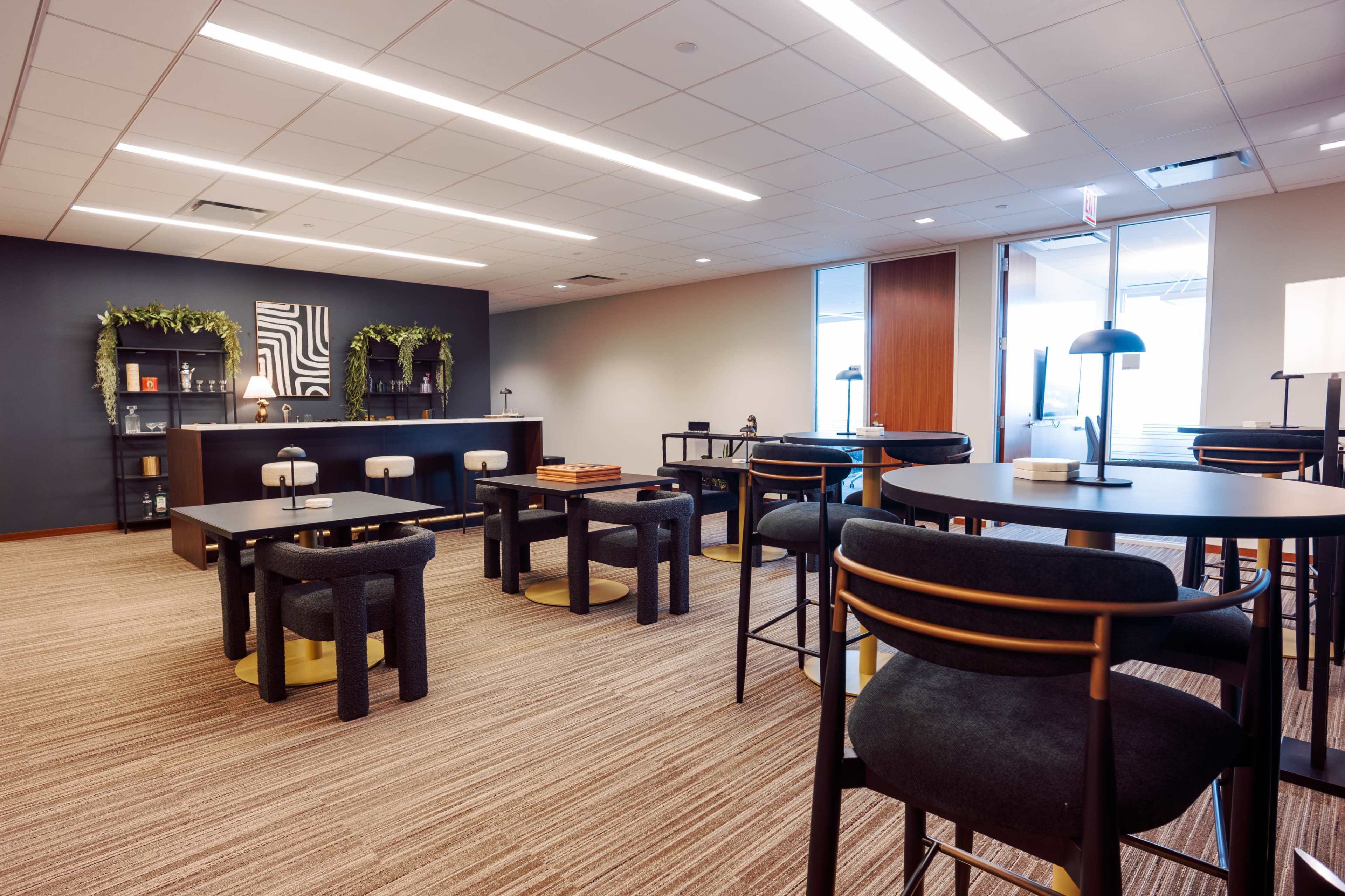 The image shows a modern office space featuring several tables, black furniture, potted plants, and a large window allowing natural light to enter.