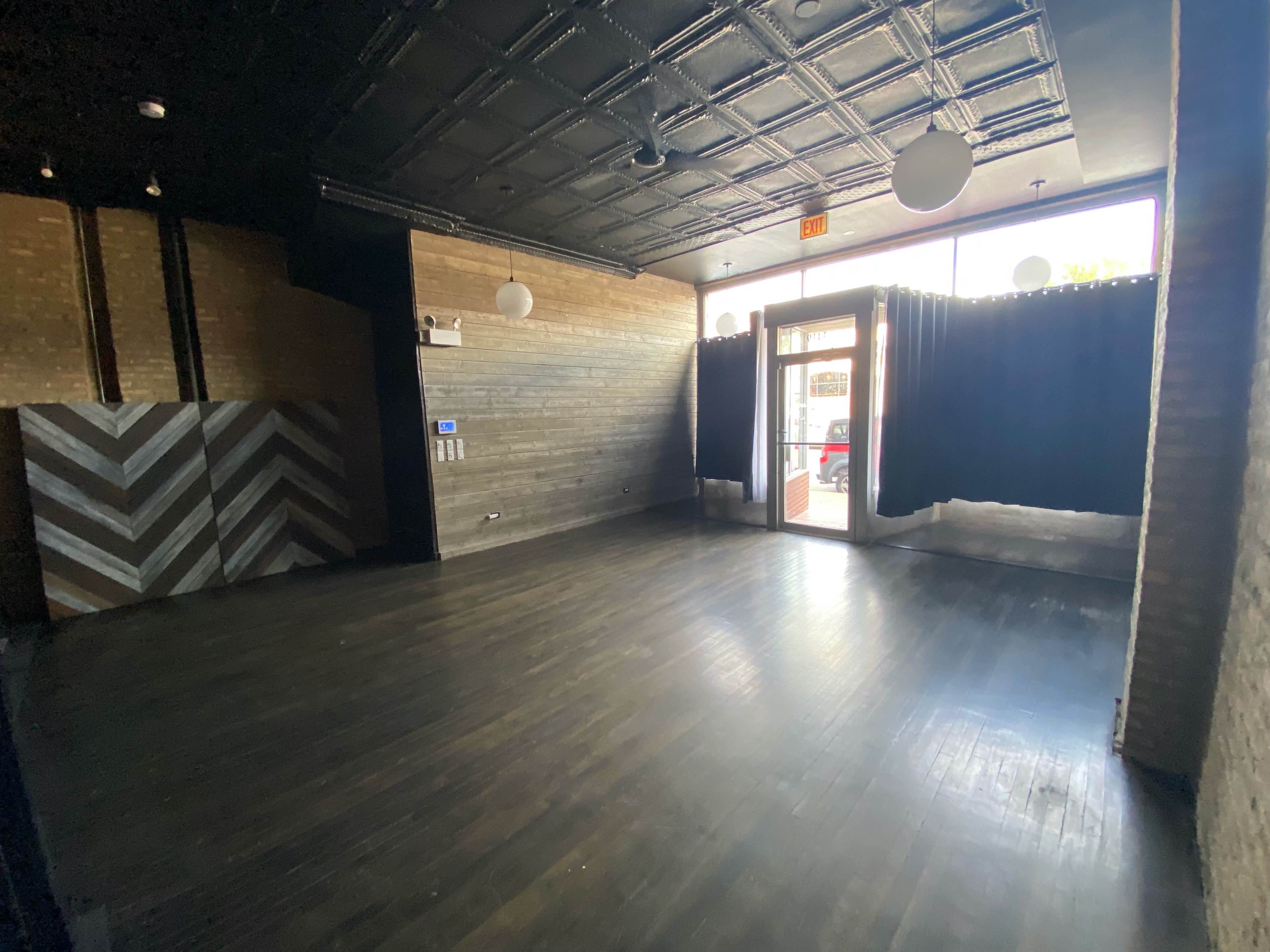 An empty indoor space with dark wooden floors, a patterned wall, and large windows covered by curtains.
