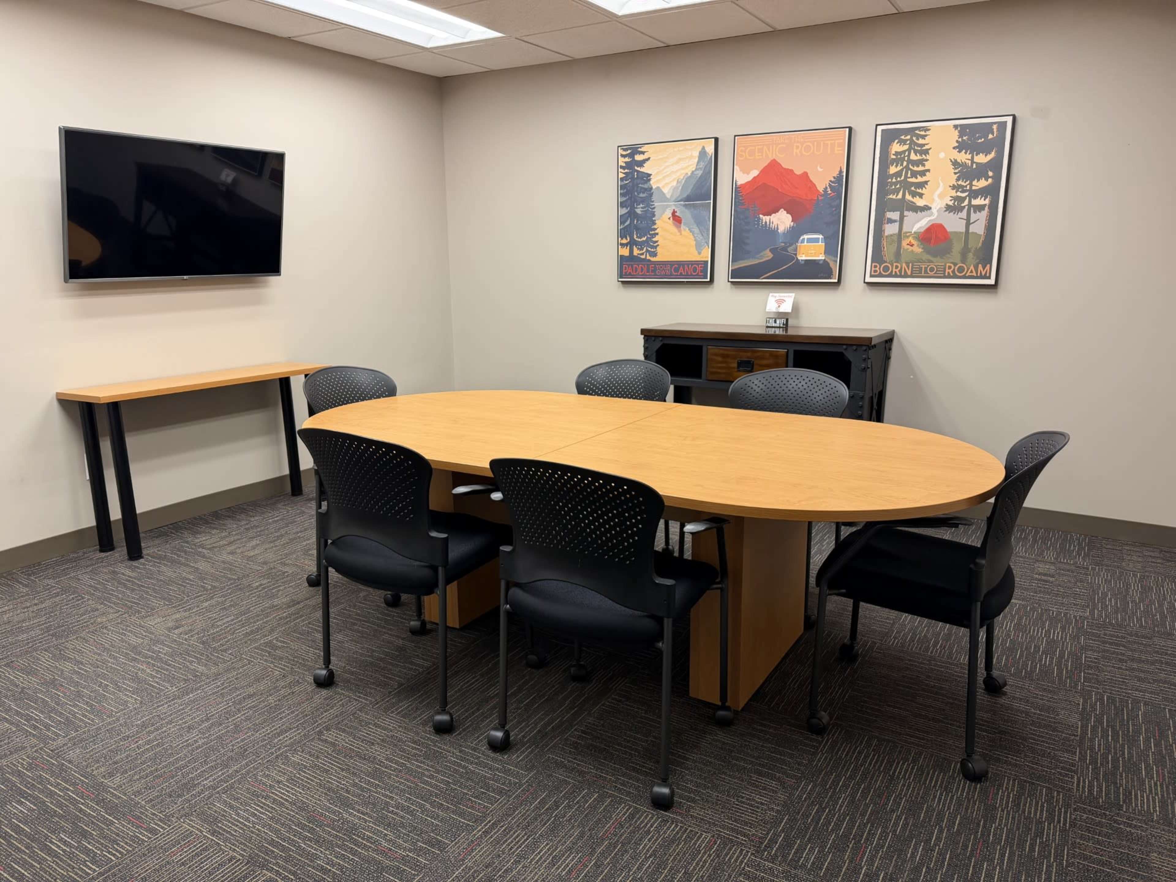 The image shows a meeting room with an oval table surrounded by black chairs, a wall-mounted TV, and two decorative posters.